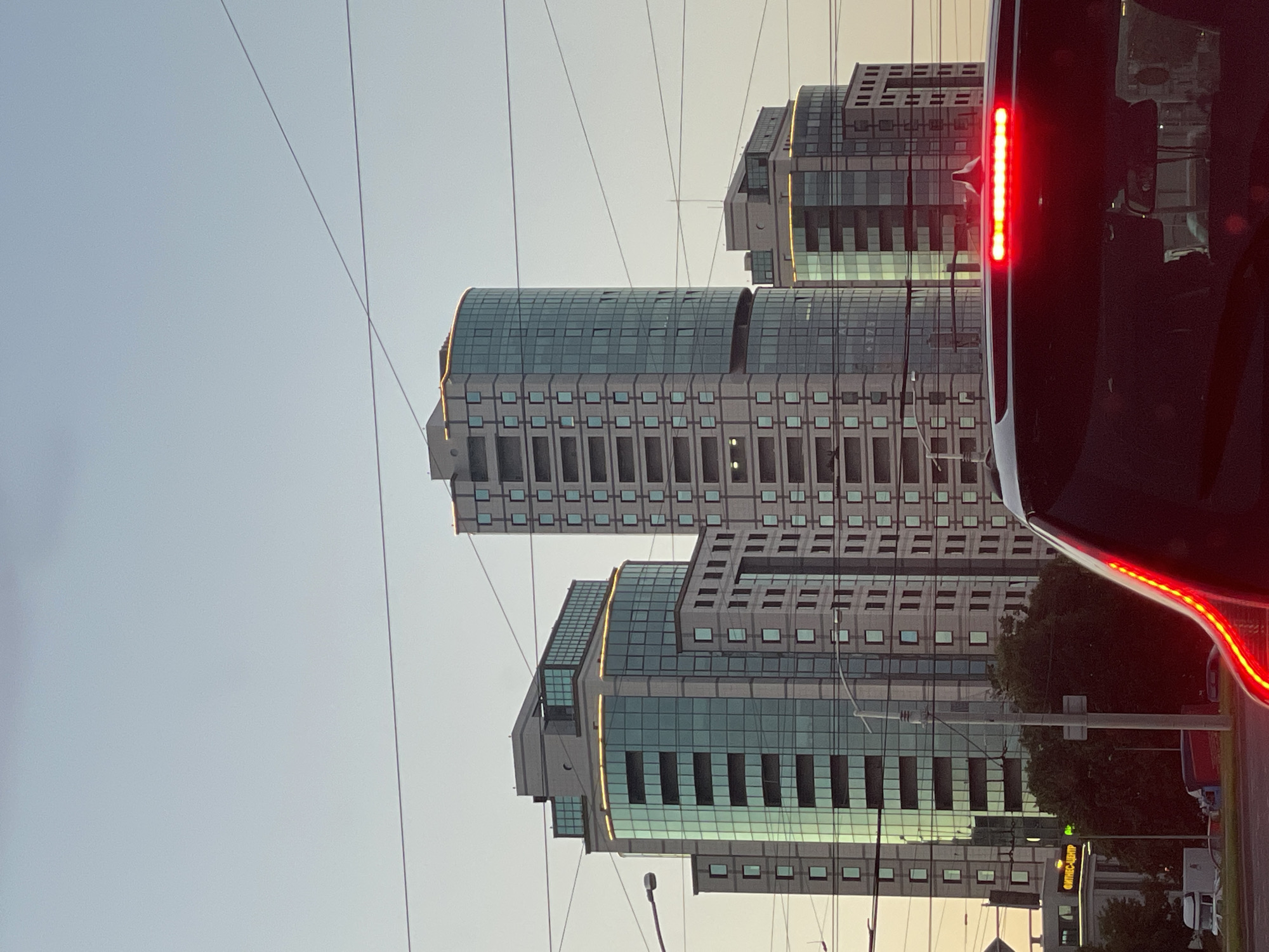 The image captures a street-level view in Minsk, Belarus, dominated by several modern high-rise buildings. The architecture features extensive glass facades, some with a teal tint, interspersed with gray concrete elements. At the top of the buildings, decorative golden lights are visible, suggesting an evening or twilight setting as the sky is a soft, pale orange.
In the foreground, the rear of a dark-colored vehicle is prominent. Its brake lights are illuminated, a bright red bar across the back, indicating it is stopped or slowing down. This vehicle is positioned such that it partially obscures the lower portion of the buildings and the surrounding environment.
The urban landscape also includes utility wires crisscrossing the sky, a common feature in many cities. A street light is visible on the left side of the frame. Trees with dark green foliage are present, hinting at a park or residential area adjacent to the business district. In the mid-ground, a building with a neon sign that reads "ФИТНЕС-ЦЕНТР" (Fitness Center) is partially visible. The overall impression is a contemporary urban scene, likely captured during commuting hours or early evening.