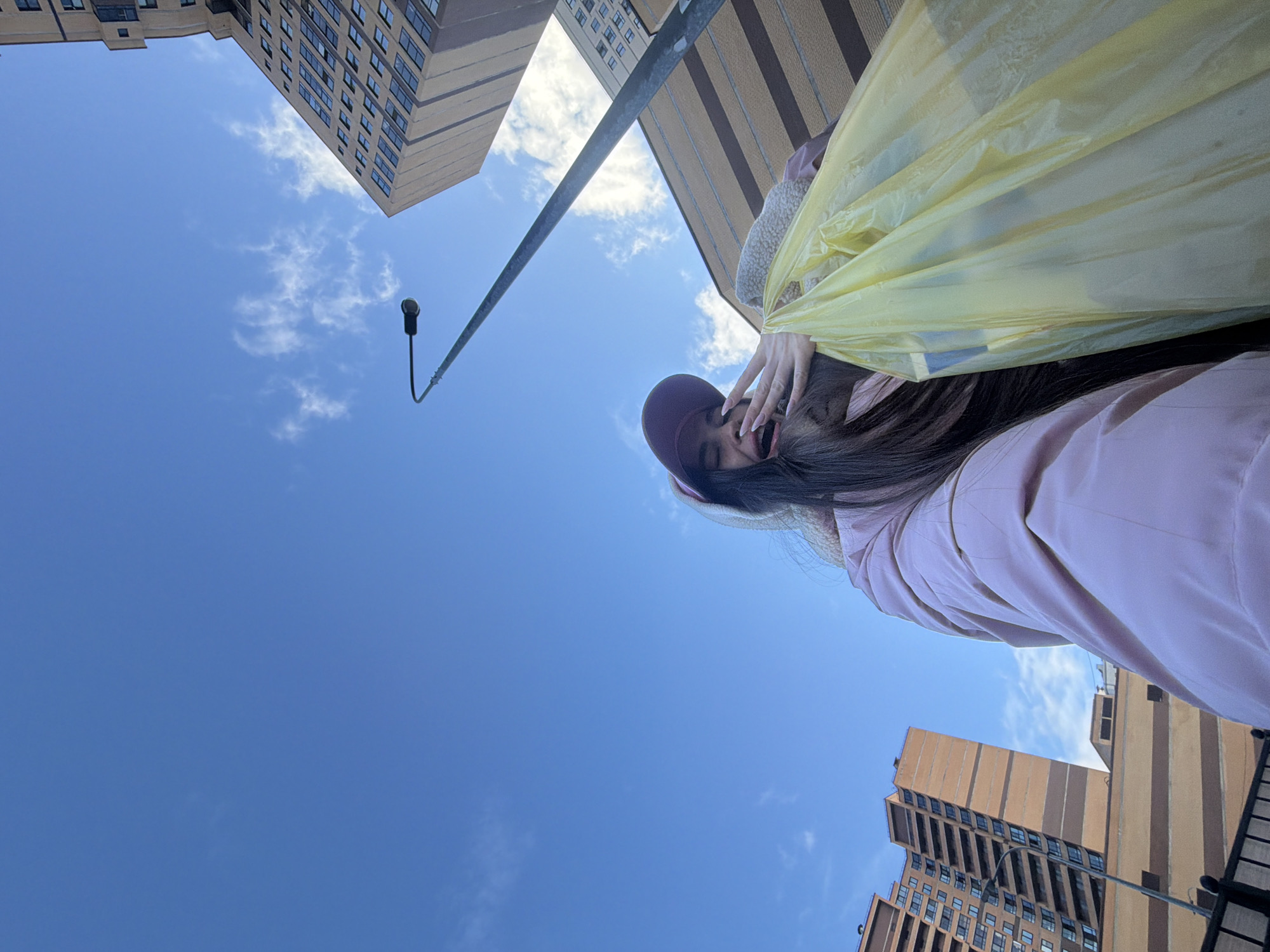 The image is taken from a low angle, looking upwards. A young woman with long dark hair and a burgundy baseball cap is partially visible in the foreground, holding a yellow plastic bag. She is wearing a light pink jacket. Her hand is near her face. Behind her are buildings, including a tall, light-colored building with numerous windows, and a building with brown and tan vertical stripes. A street light pole extends into the sky. The sky is blue with scattered clouds. The location appears to be an urban area. No evidence in the image corroborates that this is Saint Petersburg, Russia.