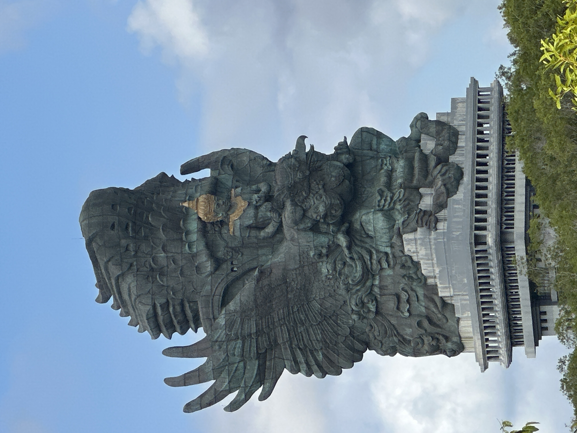 This is a vertical, medium shot photograph of the Garuda Wisnu Kencana (GWK) Cultural Park statue, located in Ungasan, Kuta Selatan, Bali, Indonesia. The statue is made of copper and brass, standing tall against a partially cloudy blue sky. The camera angle is from below, looking up at the statue's impressive scale.
The statue depicts Lord Vishnu riding on the back of a mythical bird, Garuda. Vishnu, in a seated position, wears a golden headdress and ornate jewelry. His body is a darker, greenish-bronze color, showing signs of patina from age and exposure to the elements. Garuda, the eagle-like creature, is also rendered in the same greenish-bronze material, with its massive wings spread wide. The intricate details of Garuda's feathers and Vishnu's garments are visible.
The statue rests on a multi-tiered, grey, concrete pedestal. The lower portion of the pedestal features a colonnaded design, with many rectangular columns creating a rhythmic pattern. The base of the statue and pedestal are partially obscured by lush green trees and foliage, suggesting the statue is situated in a natural or landscaped environment. The overall impression is one of grandeur and spiritual significance.