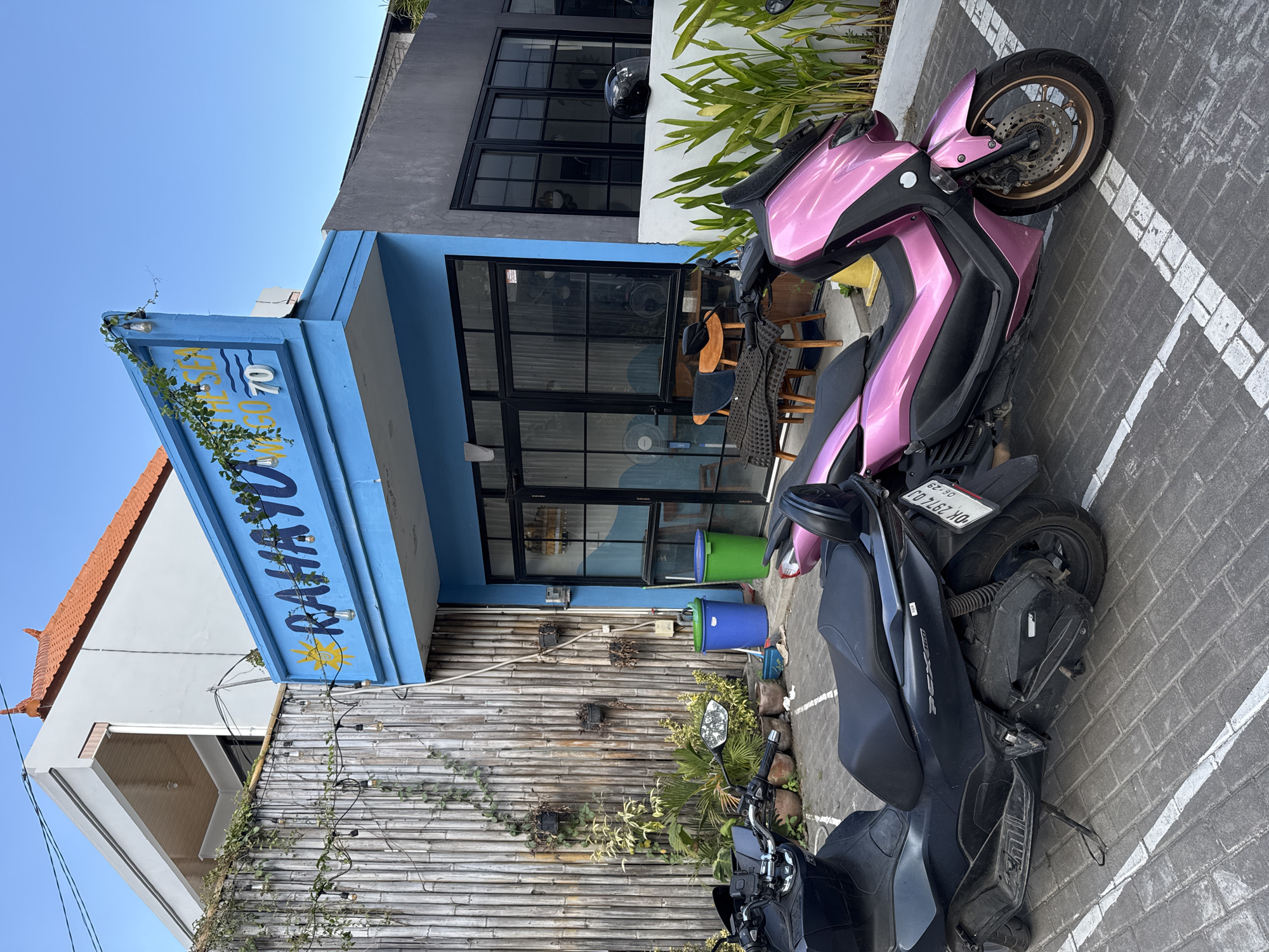 In North Kuta, Indonesia, a building with a blue facade displays the sign "RAHAYU". Adjacent to the building is a wall constructed of vertical bamboo poles, adorned with a string of light bulbs and green vines. In the foreground, two scooters are parked on a brick-patterned paved area marked with white lines. The scooter on the left is black, and the one on the right is pink. A black helmet rests on a low white wall to the right of the building, beside tall green plants. Two trash bins, one blue and one green, are visible next to the bamboo wall. The sky above is clear and blue.