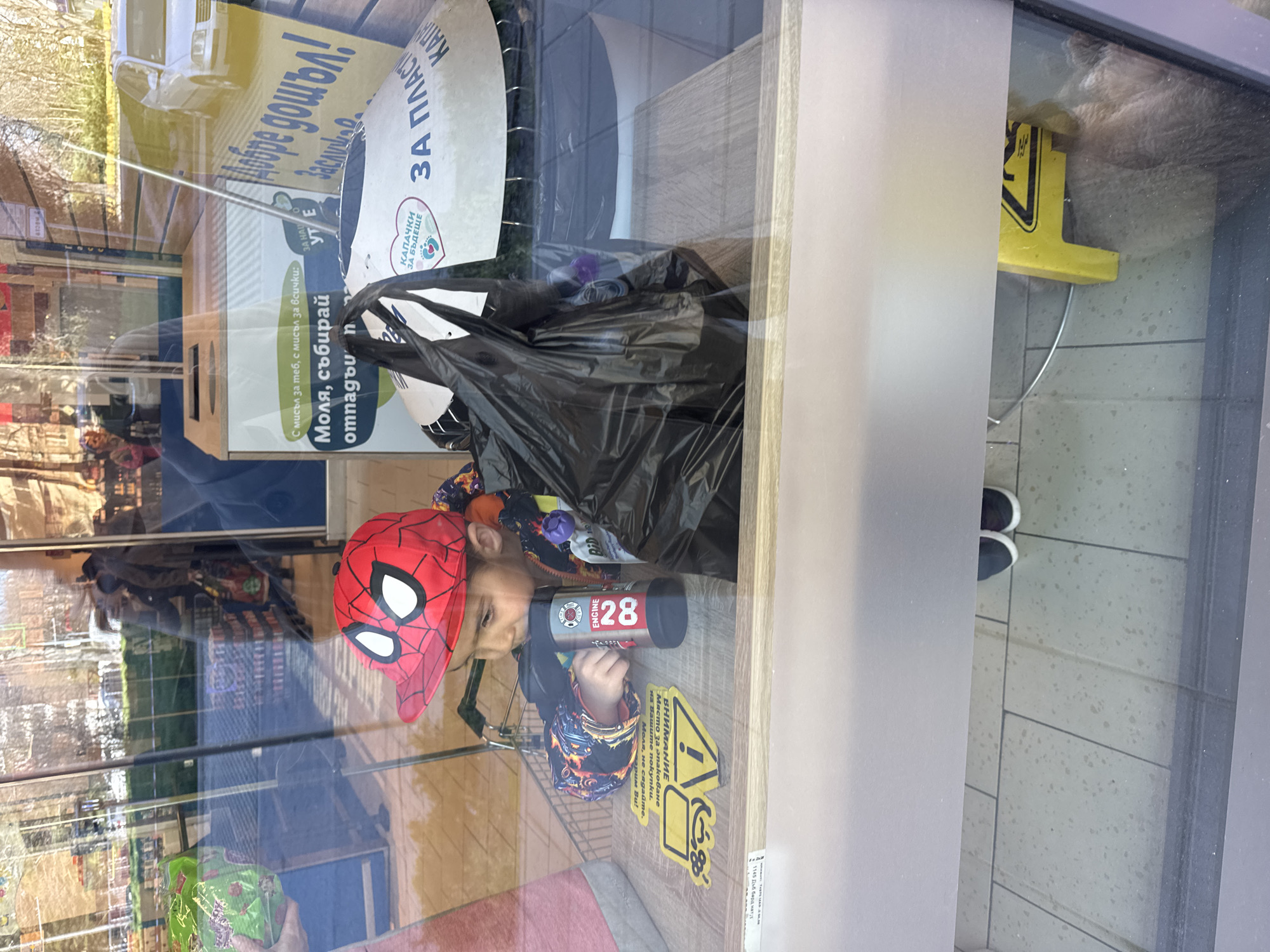 A child wearing a Spider-Man baseball cap is seen through a glass window. The child is holding a toy fire engine. The window reflects the outside environment, including trees and buildings. Inside, behind the child, is a black trash bag and a white container labelled "ЗА ПЛАСТ" (for plastic) and "КАПАЧКИ ЗА БЪДЕЩЕ" (caps for the future). Also, there is a sign saying "Моля, събирай отпадъи" (please collect waste), with the words in Bulgarian. The child is positioned near a sticker stating "ВНИМАНИЕ" (Attention) and "Място за опаковане" (Packaging location), with warnings not to sit. The image includes OCR-detected text "Добре дошъл!" (Welcome!) and a blurred reference to "1145 Дъб бард натуд." The location is most likely Burgas, Bulgaria due to the Bulgarian writing.
