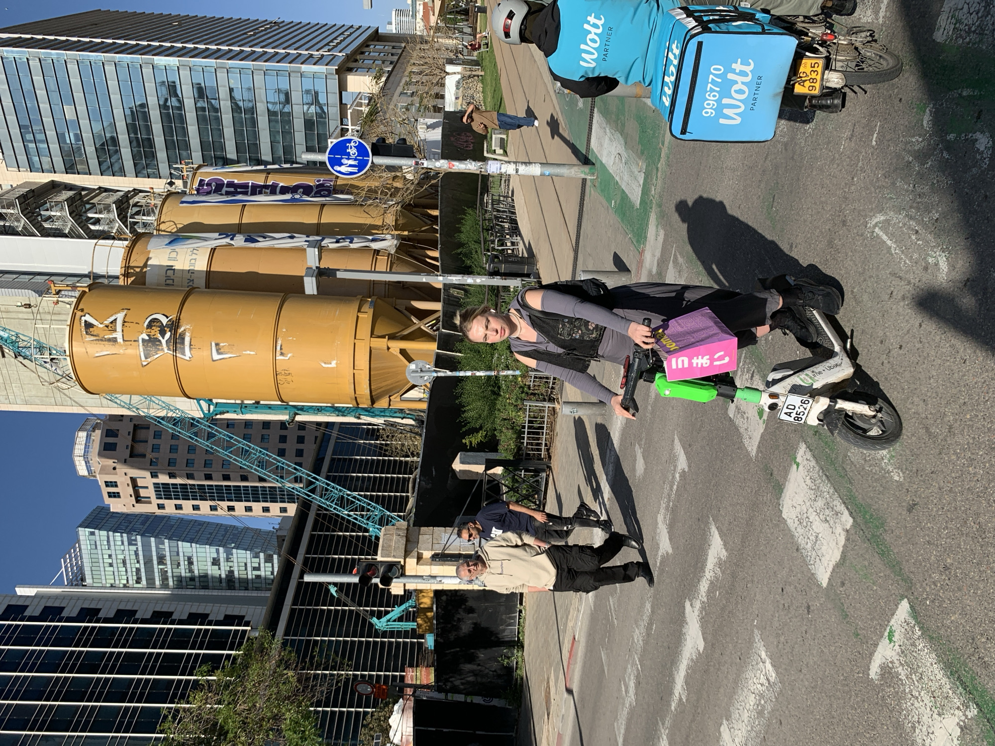 The image captures a street scene in Tel Aviv, Israel, featuring a woman on an electric scooter holding a pink bag with Japanese writing, adjacent to a Wolt delivery driver on a motorcycle. The background includes construction with large cylindrical tanks and a crane near modern high-rise buildings with reflective glass facades. Two pedestrians are walking on the sidewalk. Street signs are visible, indicating a bicycle path, and the scooter's license plate reads "AD 8526". The Wolt delivery motorcycle's license plate is "AG 9835", and the delivery bag is labeled with "996770".