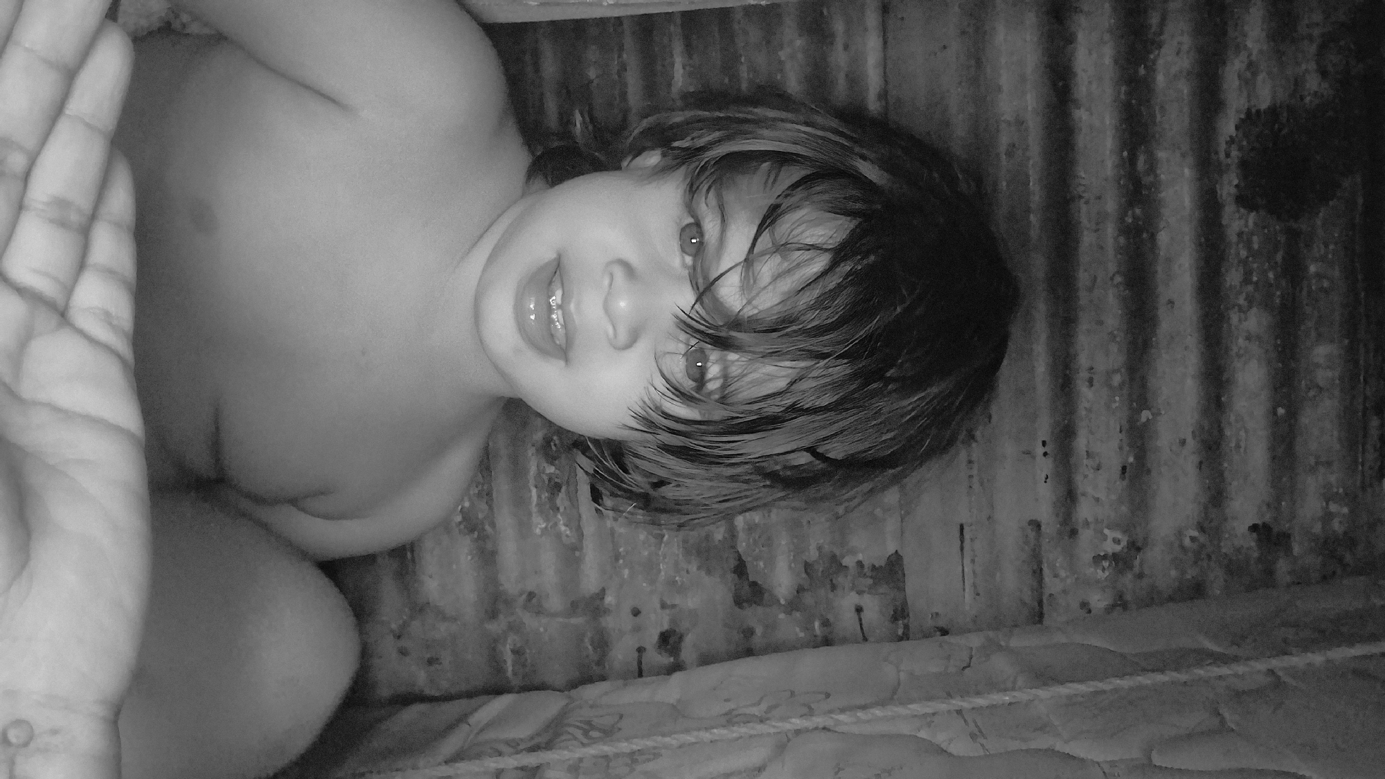 This black and white photograph depicts a young child, approximately two to five years old, looking directly into the camera. The child has dark, wet or tousled hair that falls over their forehead. Their mouth is slightly ajar, revealing upper dental braces. The child's bare shoulders and upper chest are visible, indicating they are shirtless or minimally clothed. The background consists of a textured, corrugated metal surface, likely a wall or part of a shelter, displaying signs of wear. To the right, a patterned fabric or curtain hangs vertically. A hand, belonging to an unseen individual, is visible in the lower foreground of the image. The scene is situated in the city of Vallecito, Venezuela. The lighting is low, creating a somewhat intimate and confined atmosphere.