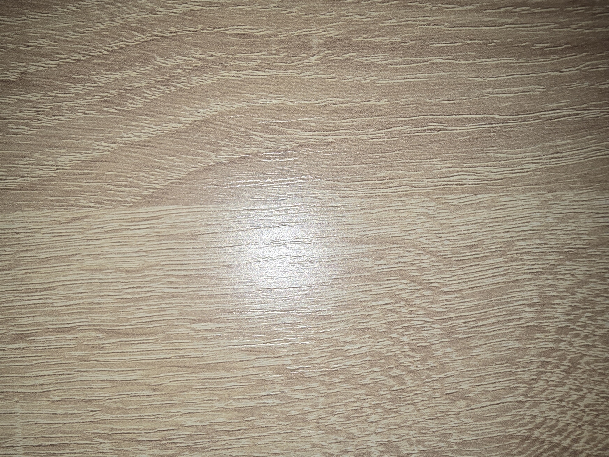 This is a close-up, textured image of a light brown wood-grain surface. The grain is characterized by thin, vertical, lighter-colored lines interspersed with darker brown lines, creating a striated pattern. There is a bright, reflective highlight running vertically through the center of the image, suggesting a glossy finish and a light source from the upper left. The texture appears to be that of laminate or a similar engineered wood product, rather than natural wood. The image does not contain any identifiable features of a city, people, or specific objects, and therefore cannot be localized to Ulan-Ude, Russia or any other location. It is purely a textural detail of a material.