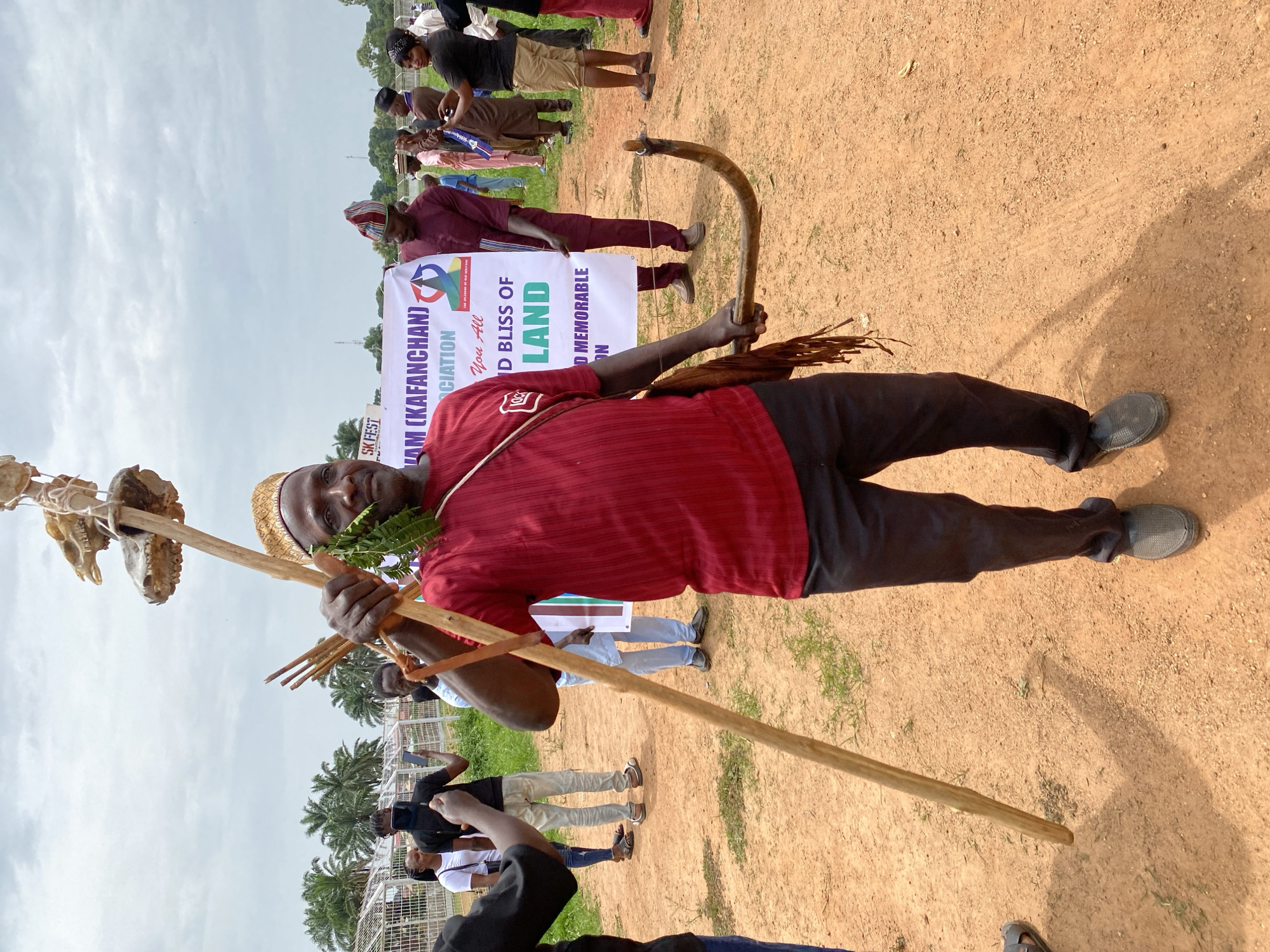 Here's a detailed description of the image:
The image depicts a man standing in an outdoor setting, possibly in Kafanchan A, Nigeria. He is dressed in a red striped shirt and black pants. He is wearing a woven hat and has green leaves around his neck. The man is holding a long wooden staff adorned with animal skulls and a bow and arrow. He has a strap across his chest.
In the background, there are other people, a banner with text "SK FEST, LAM (KAFANCHAN) CIATION, You All, ID BLISS OF LAND, D MEMORABLE ON", and trees. The ground is dirt with patches of grass. A fence-like structure is visible in the distance. People are taking pictures of the man.