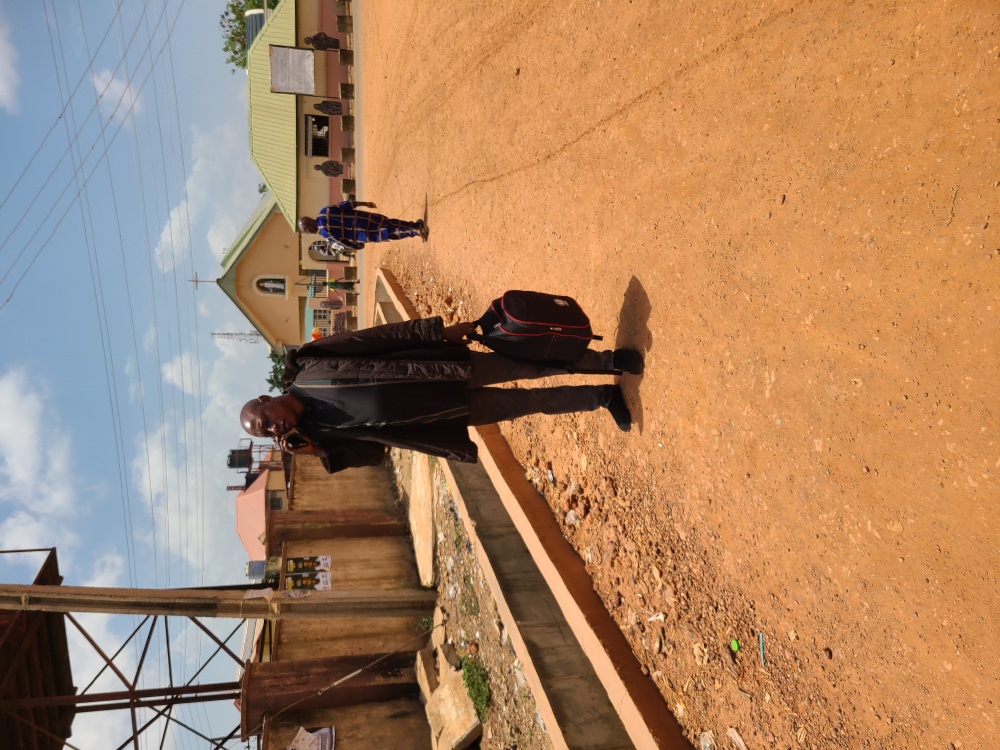 A man in a dark jacket and black shoes is walking on a reddish-brown dirt road, carrying a black and red bag. He is on the right side of the frame. To the left, there is a concrete structure with a drain, and beyond that, an open metal structure with electrical wires overhead. In the background, there are buildings, including a church with a cross on top and a light green roof. Another person is walking further down the road towards the church. The sky is blue with scattered clouds. The location is presumed to be in the city of Unknown, Unknown.
