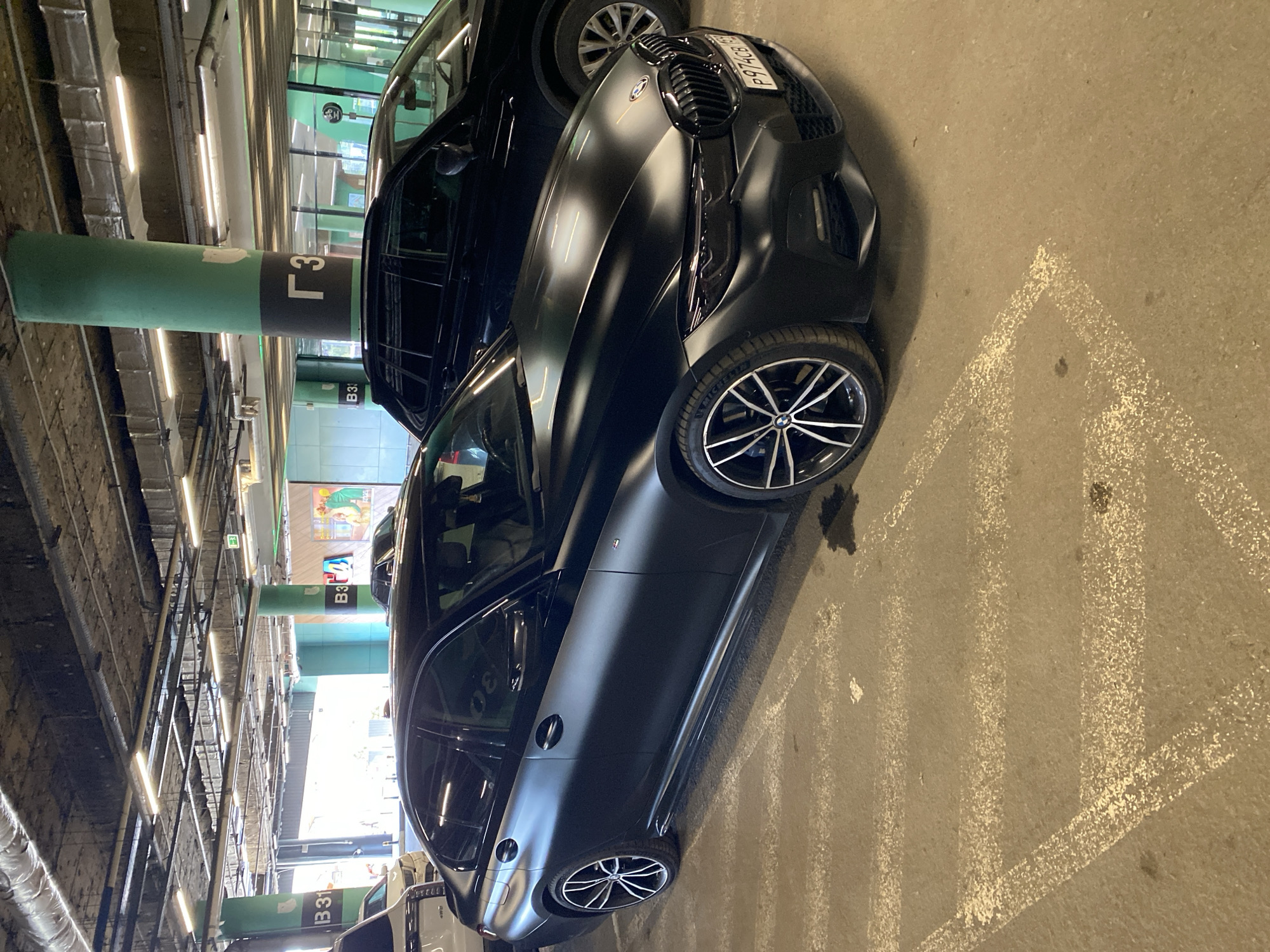 A matte black BMW i8 is parked in a dimly lit multi-story parking garage. The car is the primary subject, captured from a low angle, highlighting its sleek, futuristic design. The license plate is partially visible, reading "P97HC8". The surrounding environment consists of concrete pillars, one of which is painted green and labeled with the letter "Г" and the number "3". Another pillar in the background has "B3" visible. The ground is concrete with white painted parking lines. The lighting suggests it is either daytime with artificial lighting, or nighttime. The image is devoid of people. The scene is a typical urban parking structure.