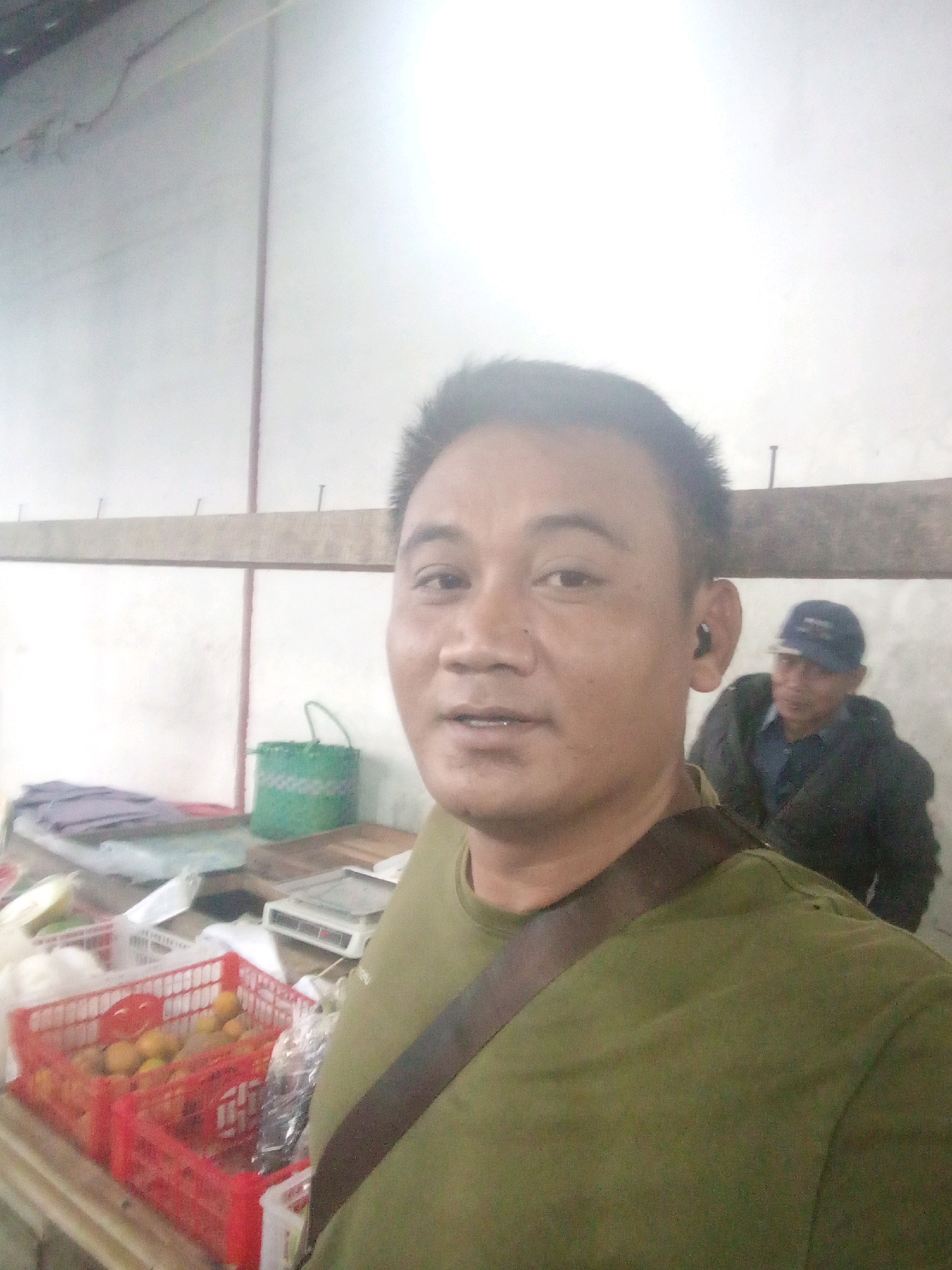 A selfie is taken by a man in a green shirt with a brown strap across his chest. He is in what appears to be a market or shop. In the background, a second man wearing a blue cap and dark jacket is visible. Various items are displayed on shelves and surfaces, including red crates filled with fruit (likely oranges or tangerines), a green woven basket, and a scale. The environment is brightly lit with plain walls. The overall impression is a candid snapshot of daily life in a local marketplace, possibly in Gentan, Indonesia, given the context provided.