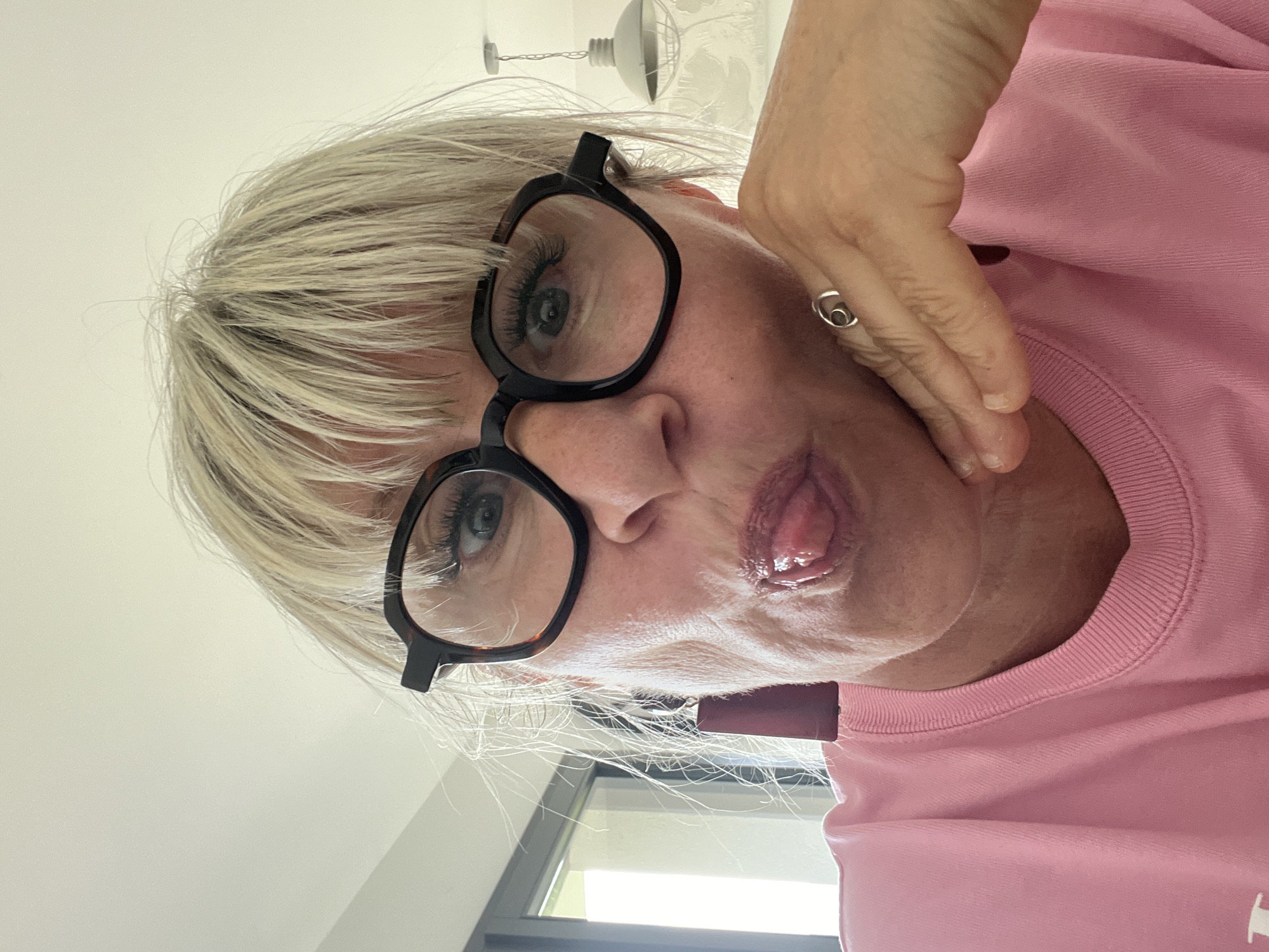 A close-up, selfie-style image depicts a woman with short, blonde hair wearing black-framed glasses. She is wearing a pink t-shirt. Her tongue is sticking out. The photo is taken at an upward angle. In the blurred background are a window and a glimpse of white ceiling and lighting fixtures. Based on image metadata, the photo was taken in Chambœuf, France.