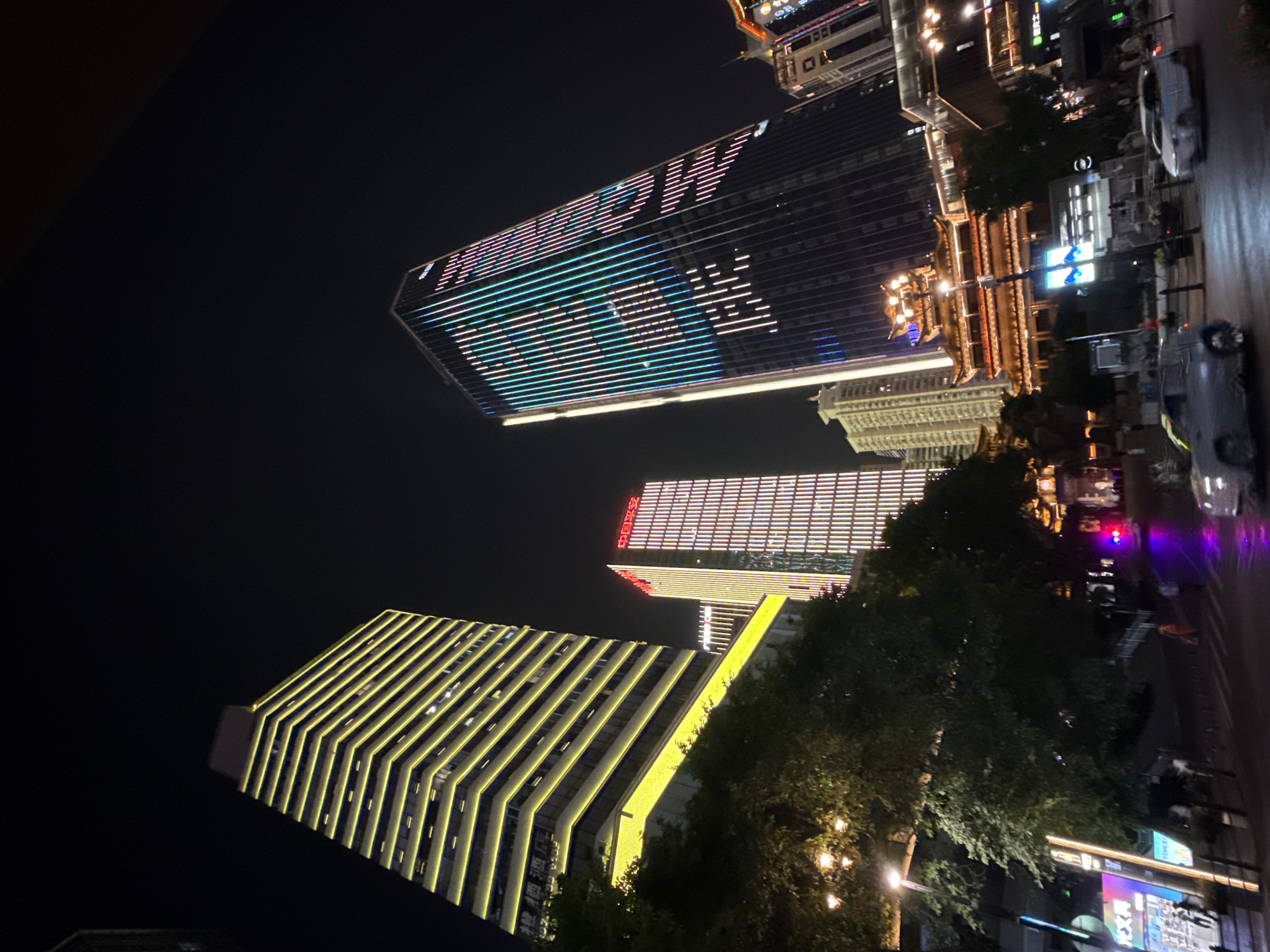 This nighttime cityscape captures a vibrant urban scene in the Yuzhong District of China. Tall buildings dominate the frame, illuminated by arrays of lights. On the left, a skyscraper is outlined with bright yellow horizontal bands. To its right, another building features vertical stripes of red and white lights, with the Chinese characters "中国平安" (China Ping An) visible at the top. Further to the right, a dark skyscraper is adorned with a dynamic LED display of multi-colored vertical lines, forming patterns of blue, green, and pink. At the base of this building, a structure with traditional Chinese architectural elements, featuring multiple tiered roofs and ornate decorations, is brightly lit. The foreground shows a street scene with cars, including a silver sedan and a white hatchback, on a wet road reflecting the city lights. Pedestrians are visible on the sidewalks, and trees with visible street lamps are interspersed throughout the scene, adding to the lively atmosphere. The sky is dark, indicating it is nighttime.