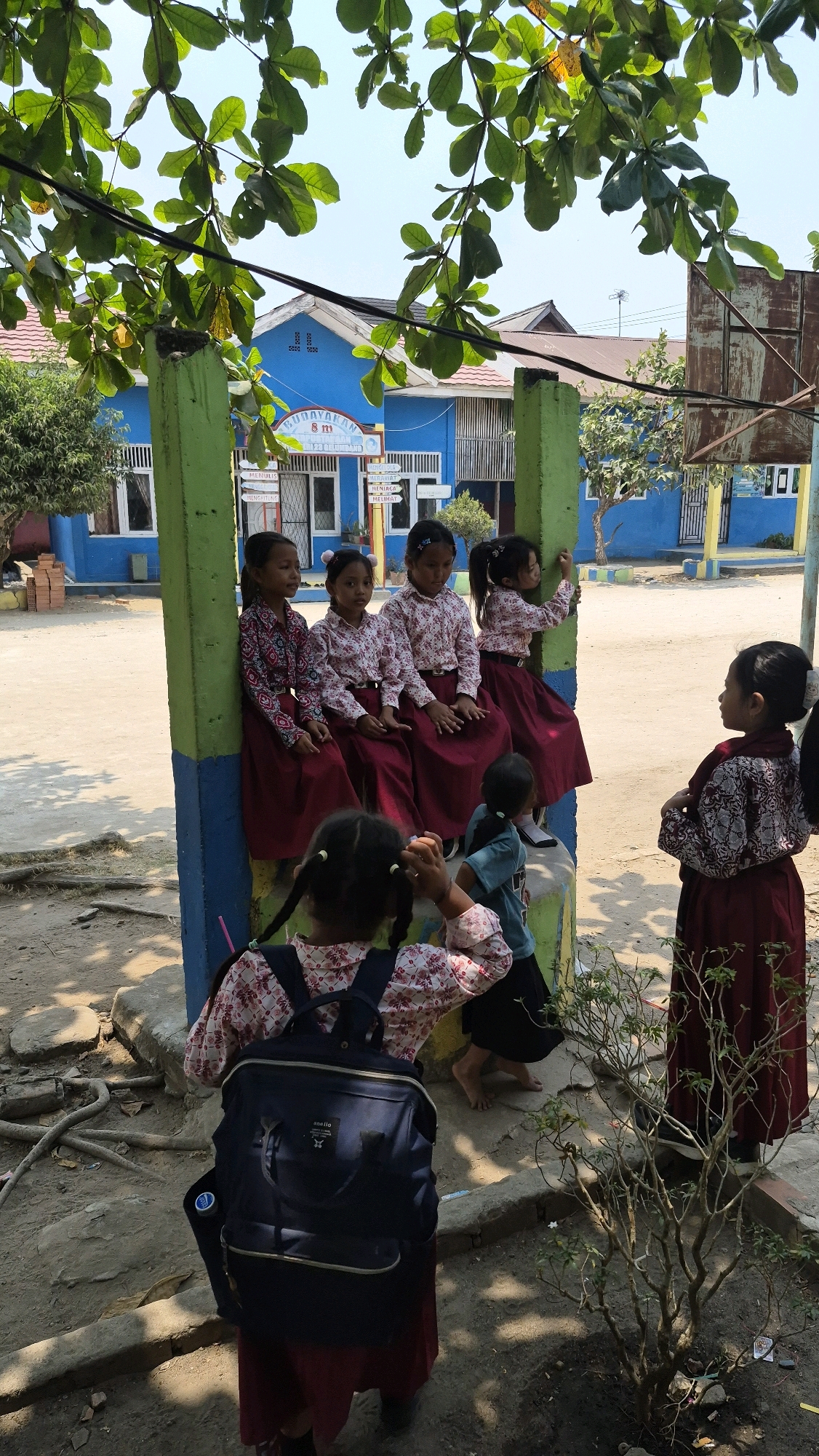 In the city of Putak, Indonesia, a group of elementary school-aged girls in maroon skirts and patterned blouses are gathered outdoors. Five girls are seated on a low, green and blue painted concrete structure, while two others stand. One girl in the foreground, seen from behind and wearing a dark blue backpack, appears to be interacting with the seated group. Another girl stands to the right, facing the seated children. The background features a bright blue school building with white trim and windows. Sunlight casts shadows on the dusty ground, suggesting it is daytime.
