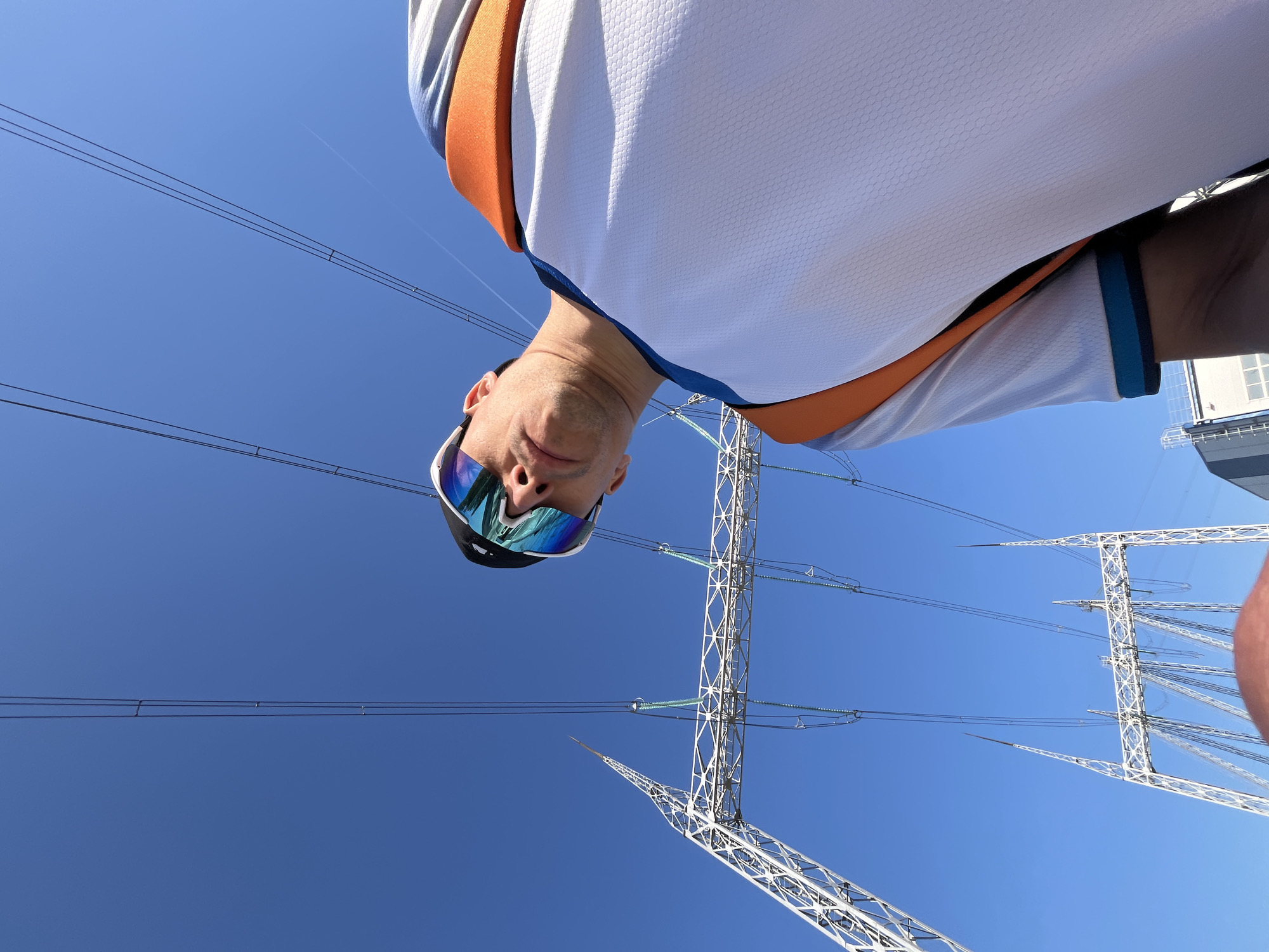 A low-angle, upward-facing image shows a man wearing sunglasses, a white shirt with orange and blue trim, and an outdoor electrical power transmission tower against a clear blue sky. The man is positioned in the upper part of the image, with the tower and power lines extending upwards and outwards. The scene appears to be in an urban or industrial area, possibly near the city of Chaikovsky, Russia, based on the request. The perspective makes the man seem to be looking down at the camera, while the tower looms overhead.