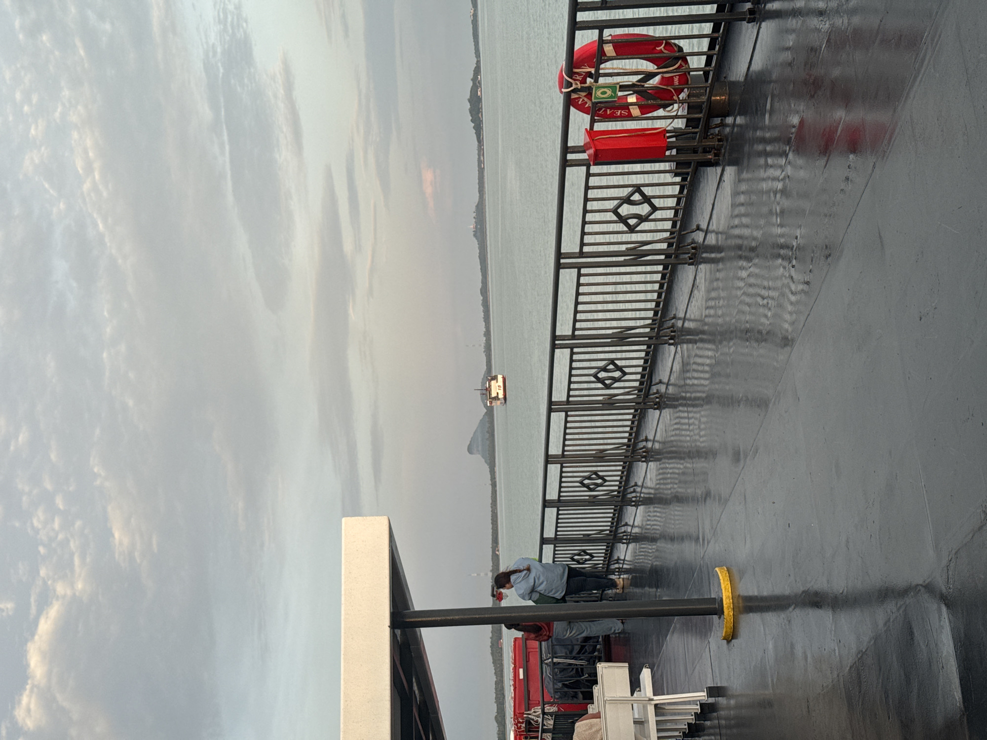 The image depicts a view from the deck of a ferry at dusk, likely near Don Sak, Thailand, given the presence of islands and a distinctive mountainous landform in the distance. The ferry's deck is wet and reflective, with visible puddles. A black metal railing runs along the edge of the deck, with a life preserver and a red box attached to it. Two individuals are standing near the railing, looking out at the water. In the background, a ferry with the number "15" visible on its side is sailing away, and islands with some structures are visible on the horizon. The sky is cloudy, with soft hues of grey and pink, suggesting either sunrise or sunset.