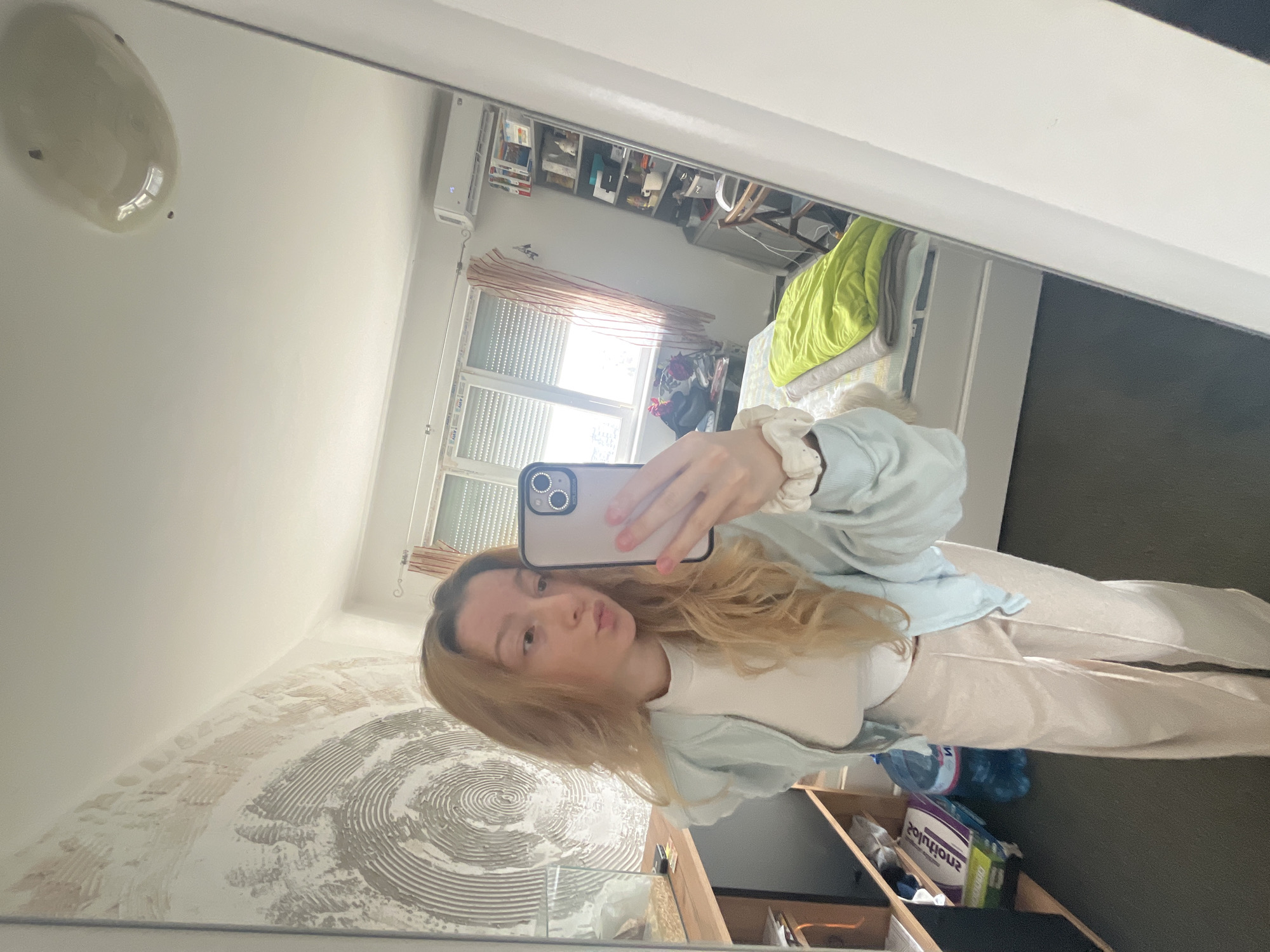 A young Caucasian female with long, wavy blonde hair captures a mirror selfie inside a residential interior in Varna, Bulgaria. She is wearing a white short-sleeved top, a light blue zipped jacket, and light beige wide-leg trousers, with a white scrunchie on her right wrist. Her lips are pursed in a slight pucker. She holds a light-colored smartphone, likely an iPhone with a black camera module, at arm's length.
The background, reflected in the mirror, shows a room with multiple features. To the subject's left, a wall exhibits a large, textured artistic application with concentric circular patterns in grey and beige tones. Below this, a wooden shelving unit contains a glass enclosure with bedding material, a blue plastic water bottle, and a white box labeled "solutions" with additional partial text. The floor is dark grey. To the subject's right, a large window with white frames and closed horizontal blinds allows daylight into the room. Striped orange and white curtains are visible beside the window. A white split-system air conditioning unit is mounted on the wall above the window, displaying "25." Adjacent to the window, a tall, multi-shelf metal unit holds various objects including books, mugs, and containers. A white-framed bed with a bright green blanket and a grey blanket is partially visible, along with a wooden folding chair. The ceiling is white and features an oval-shaped light fixture.