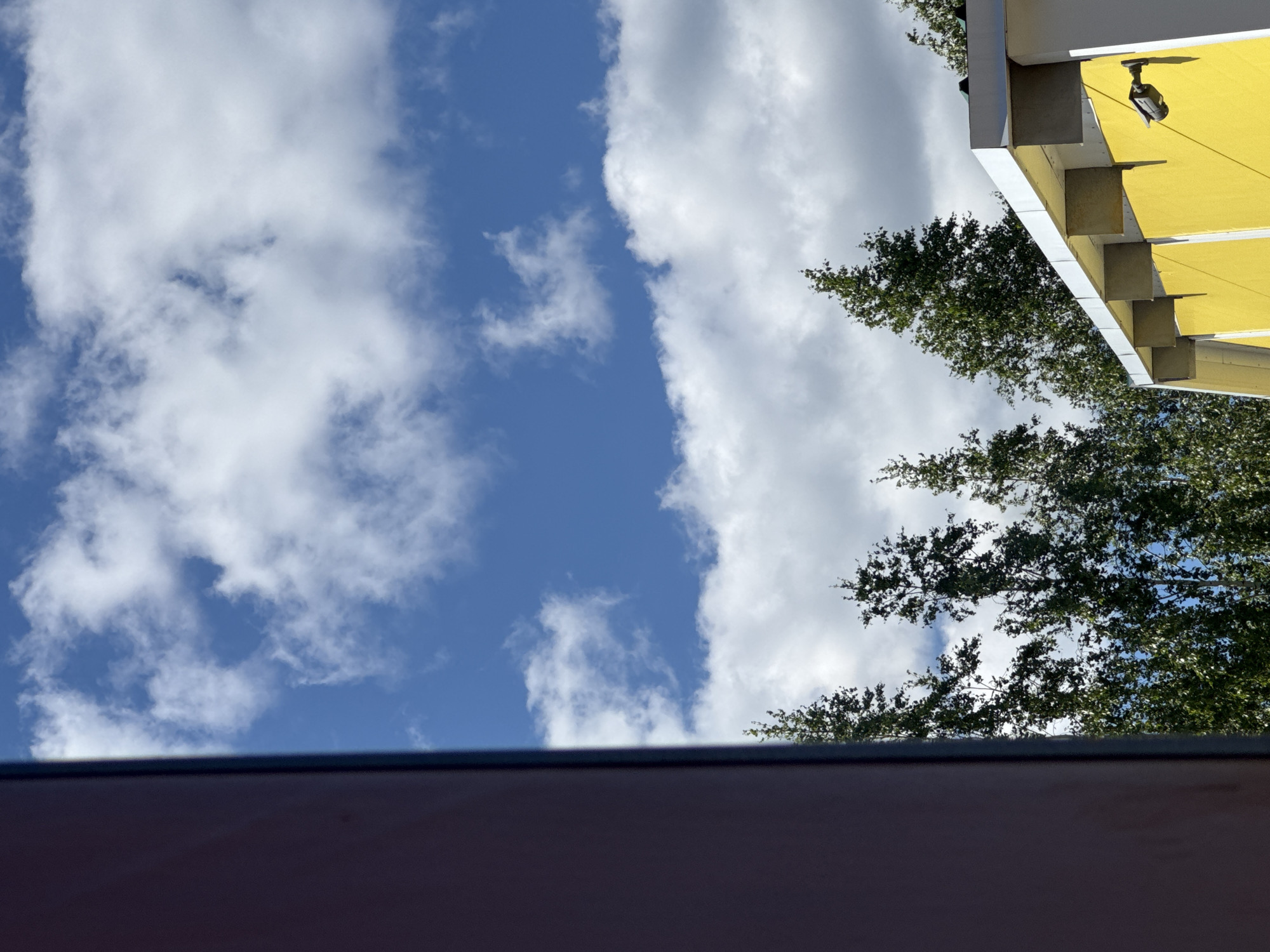 The image shows a partially obscured view of a building and trees against a blue sky with white clouds. On the left, a dark red, vertical surface occupies the foreground. To the right of this, a bright blue sky with scattered white clouds is visible. A line of tall, green trees with dense foliage forms a horizontal band across the middle of the image, separating the sky from the building below. The building's facade is painted a vibrant yellow, with a white overhang supported by grey rectangular brackets. A black security camera is mounted on the yellow wall to the right. The scene appears to be outdoors in an urban or suburban environment, possibly in the city of Zaokskiy, Russia, given the provided context.