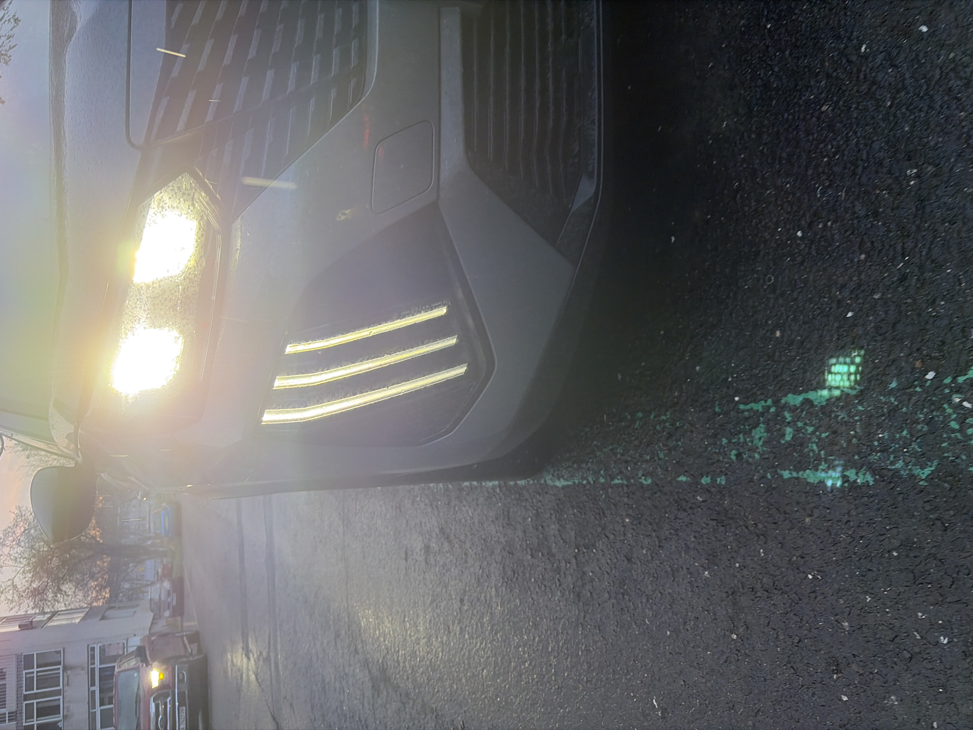 The image shows the front of a gray car with its headlights and daytime running lights illuminated. The car is parked on a dark asphalt surface. There appears to be a green painted parking line next to the car. In the background, there is a building with multiple windows. The location is Burgas, Bulgaria. There's also what appears to be a van further down the street.