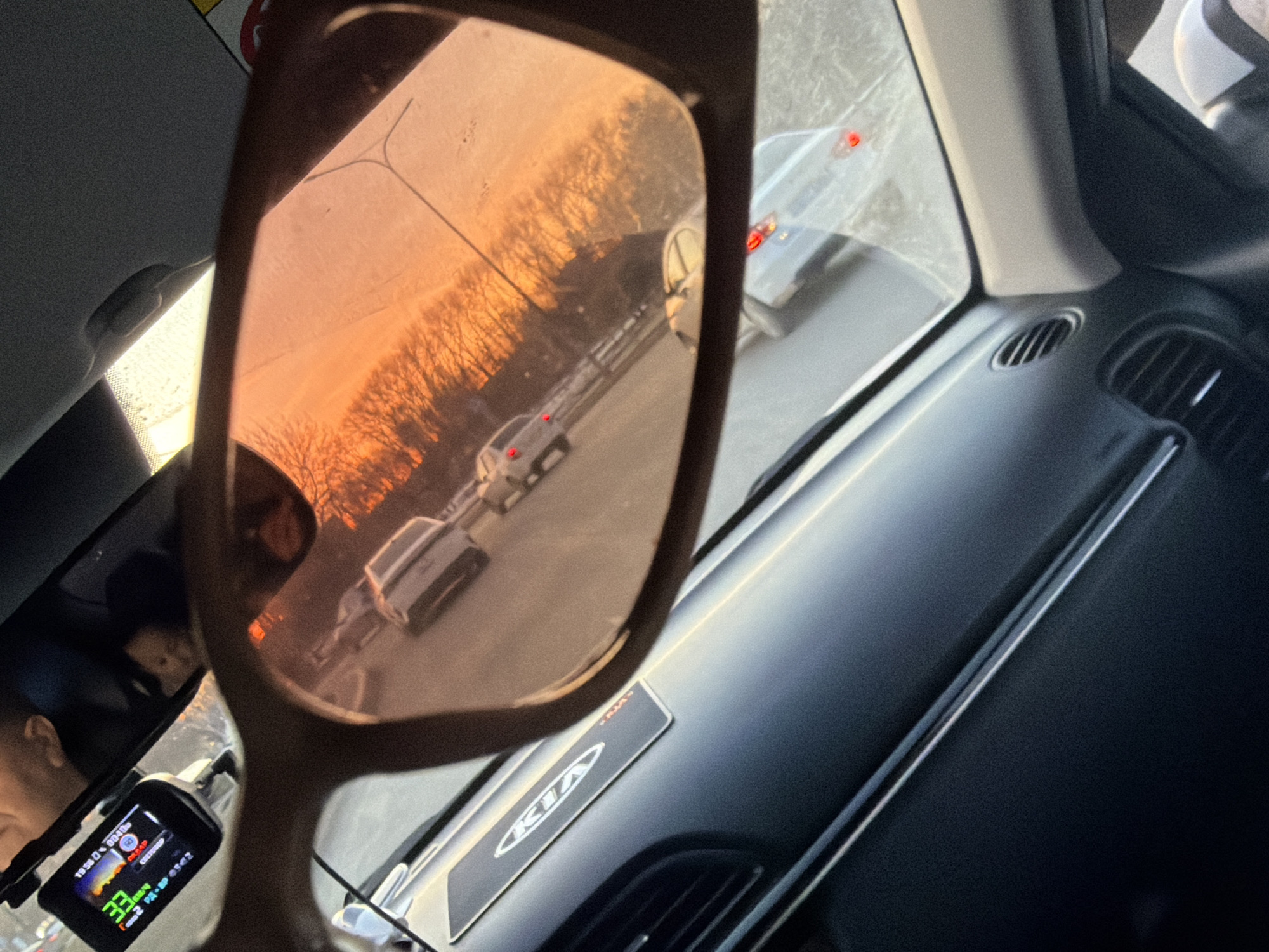 The image depicts the interior of a KIA car, specifically the dashboard and part of the driver's side. A pair of sunglasses is held up, reflecting a scene of a road at sunset with multiple cars in Perm, Russia. A dashboard camera is visible in the lower left corner, showing a speed of 33 km/h. The KIA logo is visible on the dashboard. The driver’s face can be seen in the rearview mirror. The environment outside appears to be a highway, and the sunset reflects orange hues in the sky and on the vehicles.