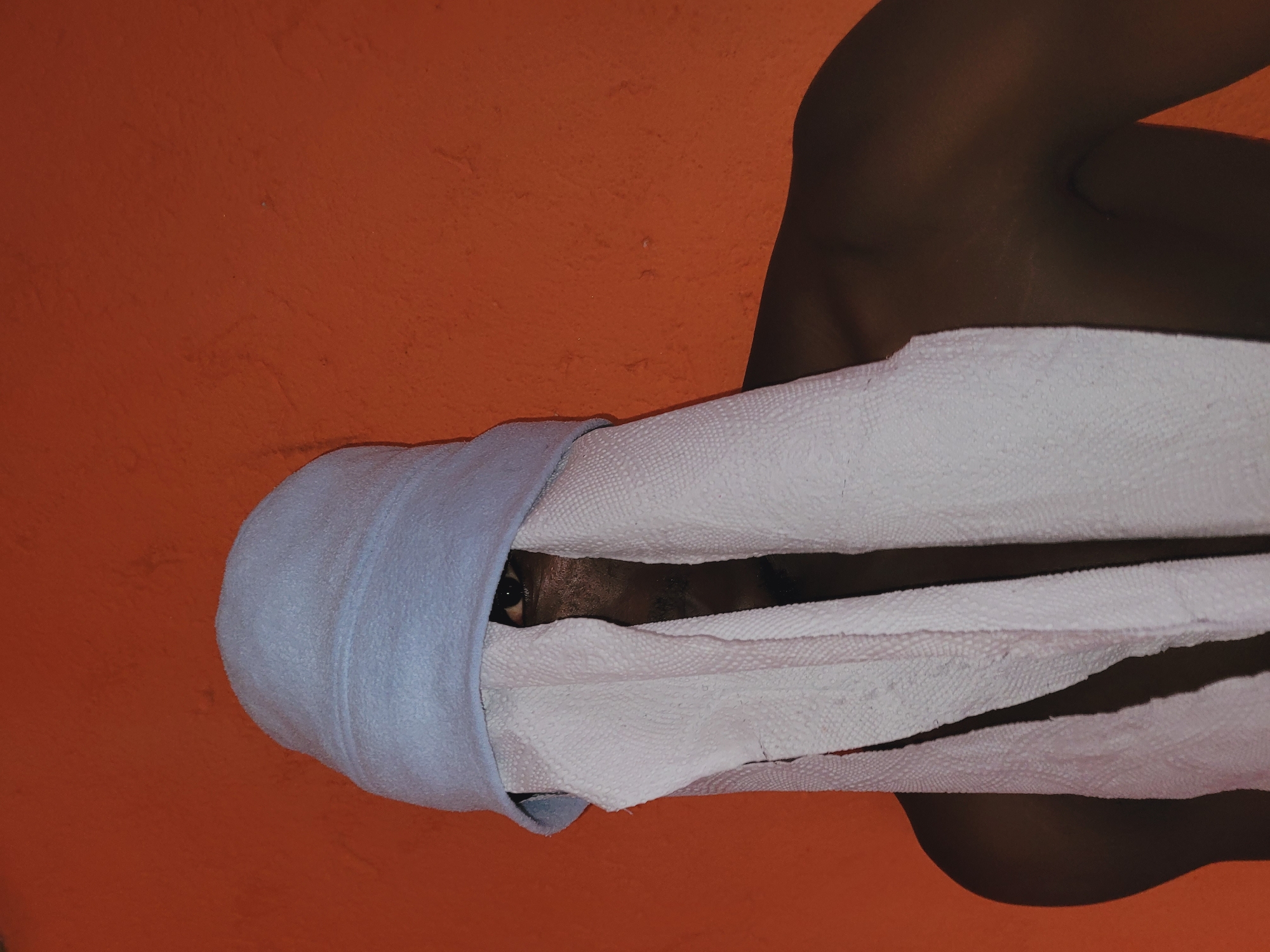 In Bukuru, Nigeria, a person with dark skin is depicted against a solid, textured orange-red wall. The individual wears a light blue beanie. White, textured strips, consistent with paper towels or similar thin fabric, hang from beneath the beanie, obscuring most of the face. A dark-colored eye is visible through a gap on the right side of the face, with a partial mustache visible below. The person's bare shoulders and upper chest are visible. The lighting is even.