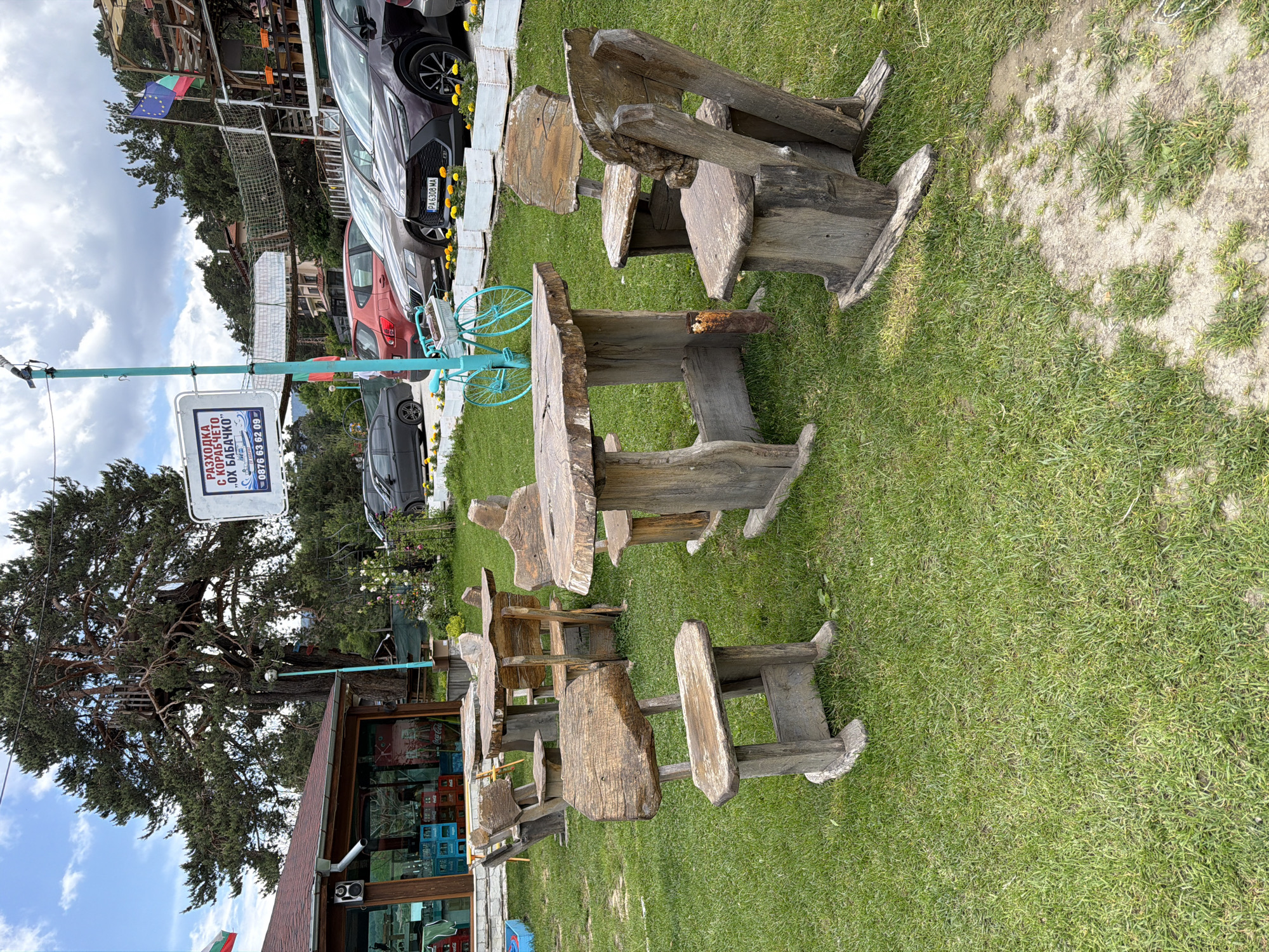 This image depicts an outdoor recreational area in Цигов чарк, Bulgaria, under a partly cloudy sky. The foreground features a grassy lawn with several rustic outdoor furniture sets, each comprising a table and chairs, all constructed from rough-hewn, natural wood slabs and logs. Patches of bare earth are visible in some areas of the lawn.
In the midground, to the left, is a single-story building with a red-tiled roof and large glass windows revealing beverages stacked inside, including Coca-Cola crates. A portion of a Bulgarian flag is visible near this building. Centrally, a tall, turquoise pole supports a white rectangular sign displaying Cyrillic text that reads "РАЗХОДКА С КОРАБЧЕТО ..ОХ БАБАЧКО" and a phone number "0876 63 62 09." A decorative turquoise bicycle with a planter basket is propped against the base of this pole. To the right of the pole, a row of four cars—a dark grey sedan, a red SUV, a light brown SUV, and a dark brown/purple SUV—are parked behind a series of white rectangular planters filled with yellow marigold-like flowers. Further to the right, behind the parked vehicles, a multi-level wooden structure with rope bridges is visible, from which a European Union flag and another Bulgarian flag are flying.
The background is dominated by a large, mature pine tree with a small wooden structure integrated into its branches on the left. Dense evergreen forests form the distant horizon line beneath a sky with scattered white clouds. No individuals are visible in the scene.