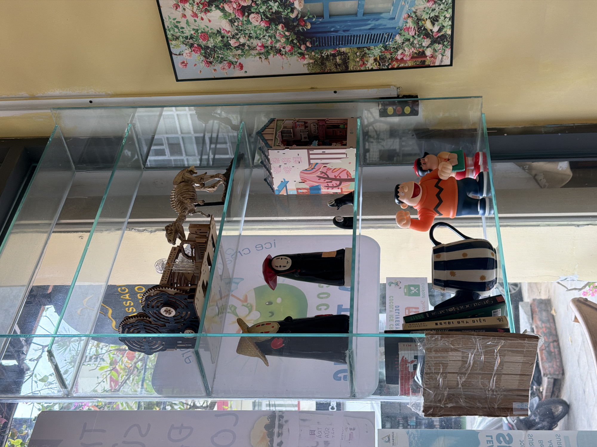 The image displays an eclectic collection of decorative items arranged within a multi-compartment glass shelf, set against a warm yellow wall. Above the shelf, a framed picture features a blue window surrounded by vibrant pink roses.
Inside the glass compartments, a variety of subjects are visible. The top-left section hosts a skeletal dinosaur model climbing a wooden structure. Adjacent to it, a detailed miniature dollhouse-like structure with Japanese text on its side occupies the top-middle. The top-right compartment contains two cartoon figures, resembling "Peanuts" characters (one in orange, one in red), next to a small panel with red, yellow, and green lights.
Further down, the middle-middle compartment showcases two "No-Face" figures from Spirited Away, one with a red hat and another with a straw hat, standing on a surface marked with "Ice C" and "GOG." The middle-right compartment features a blue and white striped mug with stars, stacked upon several books. Identifiable titles include "SHERLOCK HOLMES," a book by "HOSSEINI" titled "NGƯỜI ĐỊA DIỄN" (Vietnamese for "The Kite Runner"), and another by "MARISSA MEYER." Below these books lies a cardboard package wrapped in plastic.
The setting appears to be indoors, likely a cafe or shop, characterized by its decorative elements and warm lighting. No specific activity or event is taking place; the image is a still life. The bright, natural light suggests it is daytime. Location cues are confirmed by visible Vietnamese text such as "NGƯỜI ĐỊA DIỄN" and "COCOPA ICE CB," aligning with the provided context of Bac Nha Trang Ward, Vietnam. Other visible texts include "SSAG," "JOJ," "ÁSIT," "TIM," "ΓΕ," and various numbers. The diverse collection of whimsical objects creates a personal and inviting atmosphere.