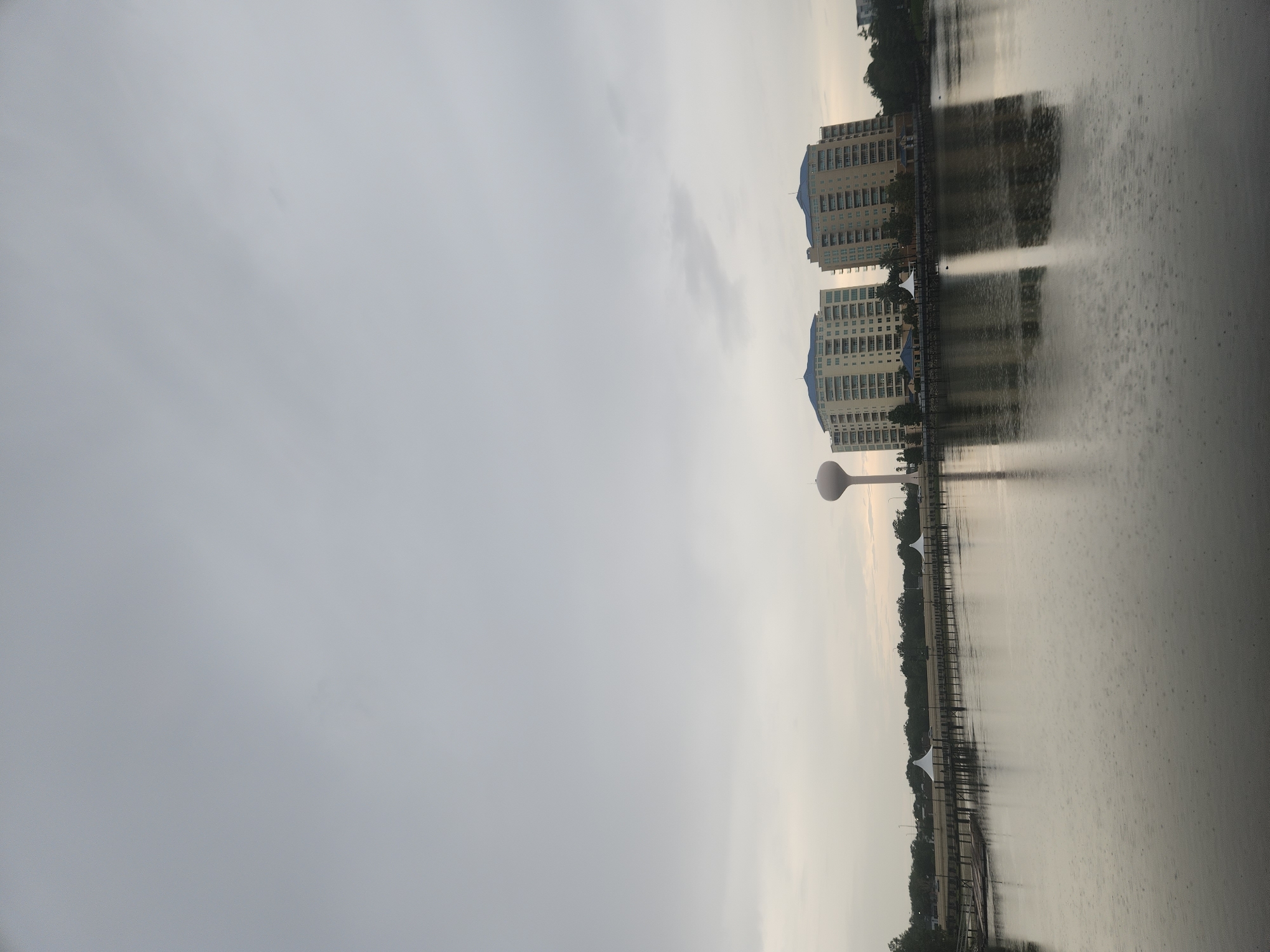 The image depicts a serene water scene in Altamonte Springs, United States of America, under an overcast sky that suggests impending rain. The foreground is dominated by a calm body of water, reflecting the muted light and the gentle ripples caused by raindrops. A wooden pier extends into the water from the left side of the frame, lined with pilings and wooden planks, leading towards structures on the opposite bank.
In the middle ground, two identical, multi-story apartment buildings with blue roofs stand prominently. To the left of these buildings, a tall, cylindrical water tower with a domed top is visible. The buildings and the water tower are reflected in the water, their forms slightly distorted by the surface disturbances.
The background consists of a tree line and what appears to be a road or walkway behind the pier, with several white, conical tents spaced along it. The overall atmosphere is tranquil and slightly somber, characteristic of a pre-rain or light rain day. There are no people or animals clearly visible in the frame. The scene is captured from a vantage point that overlooks the water and the cityscape beyond.