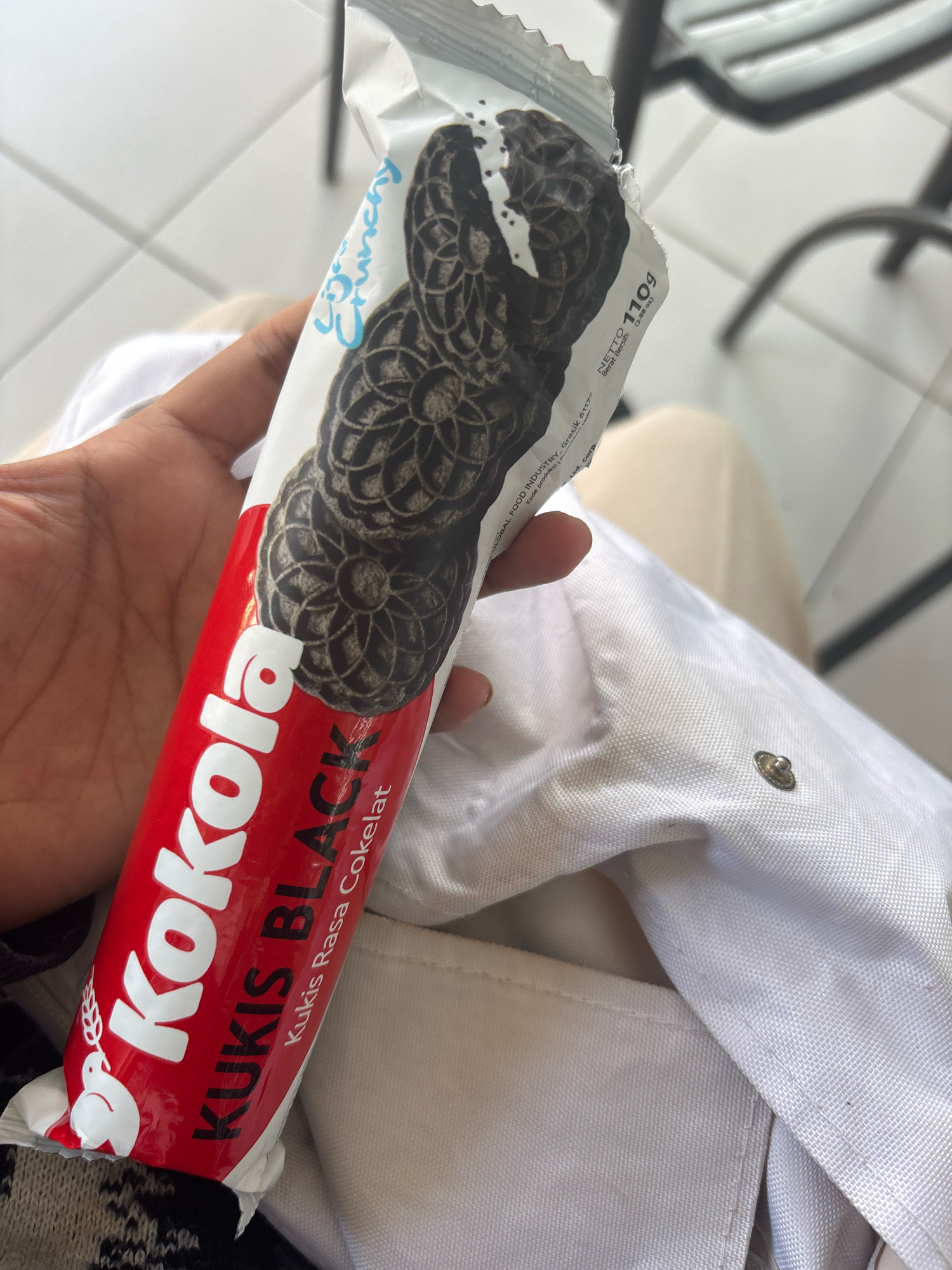 A person's hand holds a red and white packaged roll of Kokola Kukis Black chocolate cookies. The packaging features white text and graphics against a red background on the lower portion, and white with black cookie images on the upper portion. The upper part of the packaging also displays the word "Crunchy" in blue script. The cookies appear dark brown with cream filling. The person's hand is a light brown skin tone and is positioned to the left of the cookie packaging. The background is blurred, showing a light-colored tiled surface and some dark metal furniture, suggesting an indoor setting likely in a commercial or public area. The lighting is bright and natural.