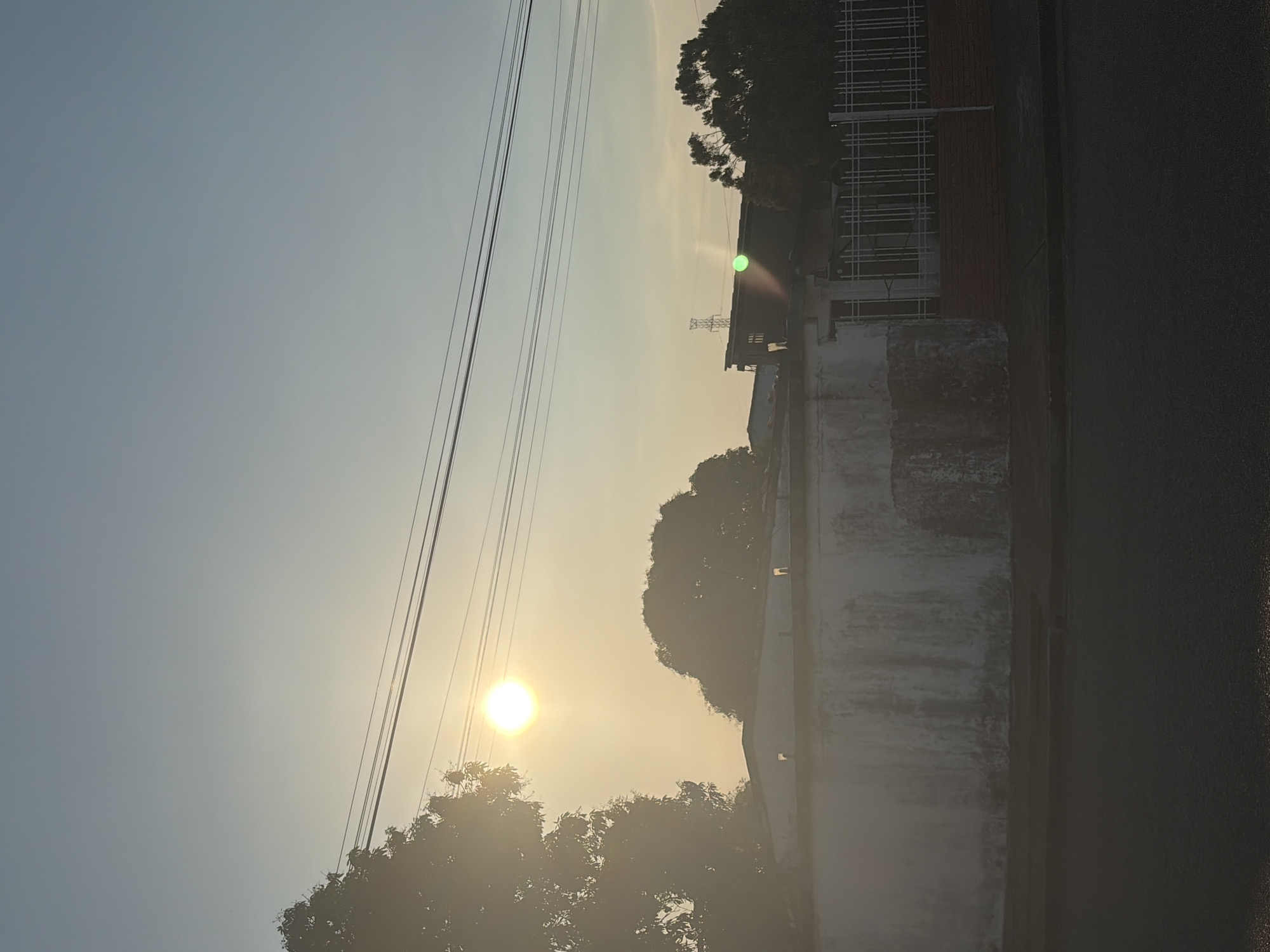 The image shows a street scene in Guayana City, Venezuela during daytime, probably late afternoon or early morning based on the position of the sun. A large concrete wall runs along the right side of the image, adjacent to a paved road. Beyond the wall, glimpses of buildings with metal scaffolding and foliage from trees are visible. Overhead, power lines stretch across the sky. The sun is visible through the trees, creating a hazy effect.