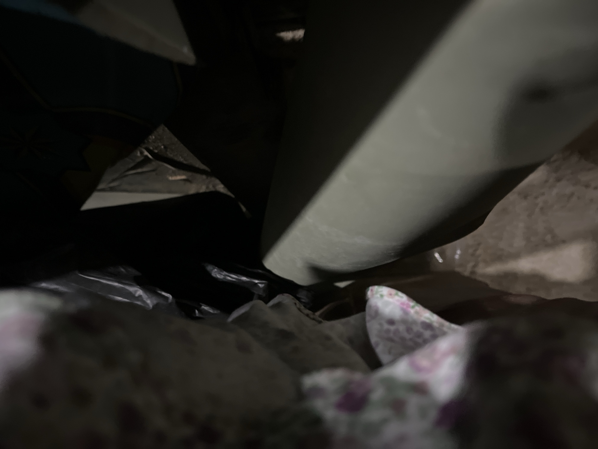 The image shows a close-up, low-angle view of the interior of a dark space, possibly a bag or container. A large, smooth, gray cylindrical object dominates the upper right portion of the frame, casting shadows. The lower portion of the image is filled with crumpled, metallic-looking material and what appears to be fabric with a floral pattern in shades of pink and white. The overall lighting is dim, with some areas obscured by shadow. The scene is static and does not depict any discernible action or interaction. There are no identifiable people or specific landmarks that would allow for a definitive location in Benuaraja, Indonesia.