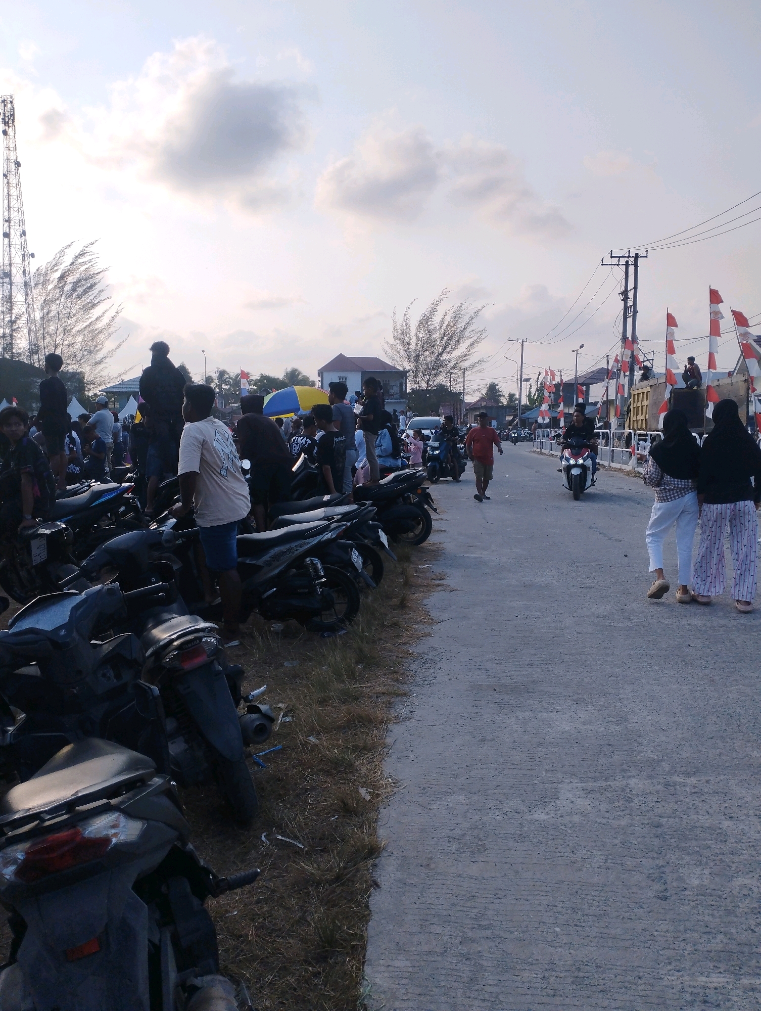 In Kuala Samboja, Indonesia, a crowd gathers near a road lined with parked motorcycles and scattered debris. A cell tower stands prominently to the left, with dry grass and vegetation bordering the roadside. People are seen standing, sitting on motorcycles, and walking along the street. Red and white flags are displayed along the right side of the road, suggesting a festive or nationalistic event. The sky is partly cloudy, with a muted light indicating it is either early morning or late afternoon. Buildings and trees are visible in the background, contributing to the urban or semi-urban setting.