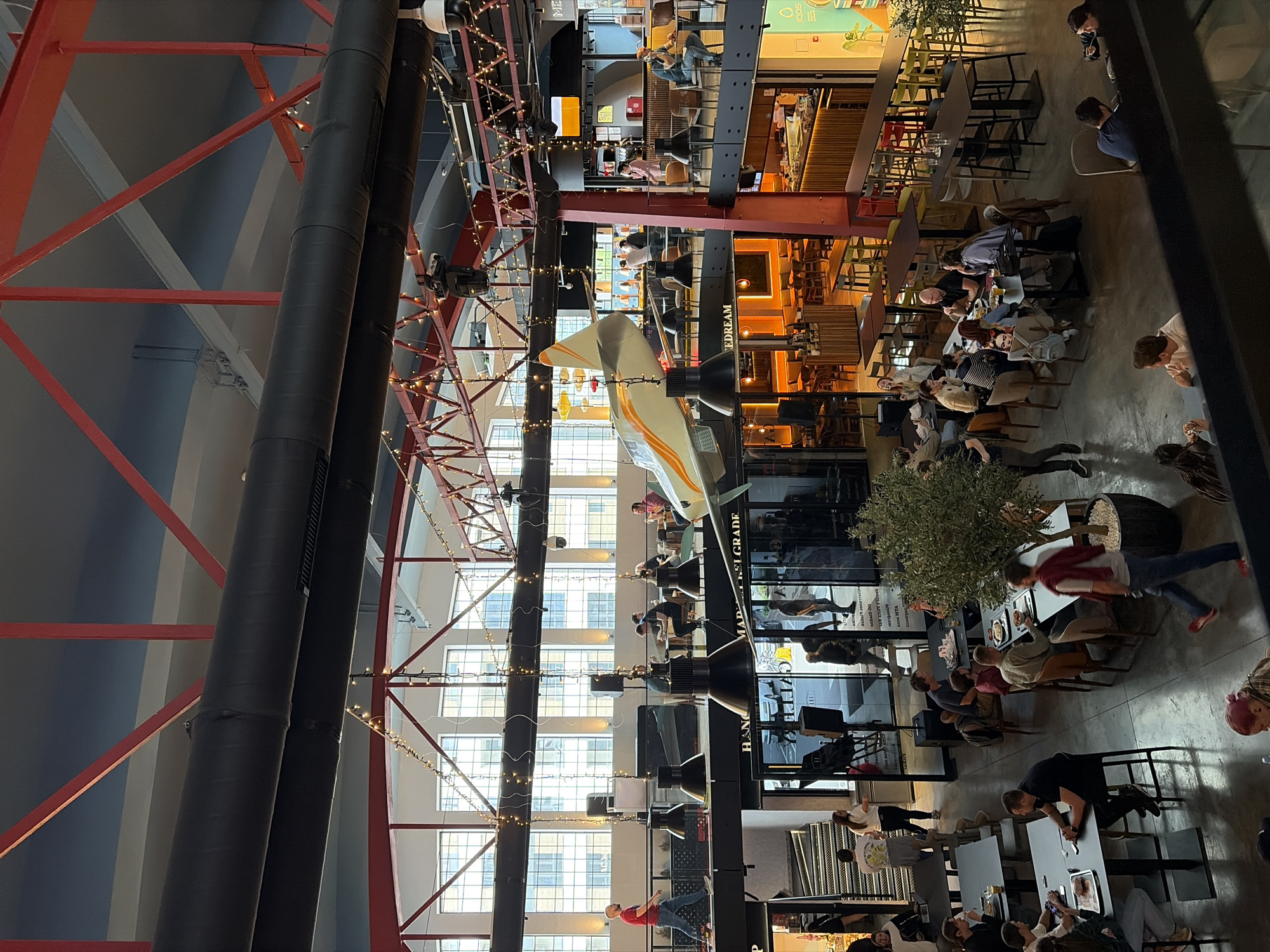 The image depicts the interior of a large, modern space, possibly a mall or a food court in Belgrade, Serbia. The architecture features a high ceiling with exposed red steel beams and large black pipes. Light fixtures are strung across the ceiling, adding to the ambience.
The area is populated with people seated at tables, suggesting a dining or gathering place. They are engaged in various activities such as eating, drinking, and socializing. The tables and chairs are arranged in multiple rows. Some tables are occupied by groups, while others are occupied by individuals.
A notable feature is a large aircraft model suspended from the ceiling, attracting attention and serving as a decorative element. The background features a multi-story structure with windows providing natural light.