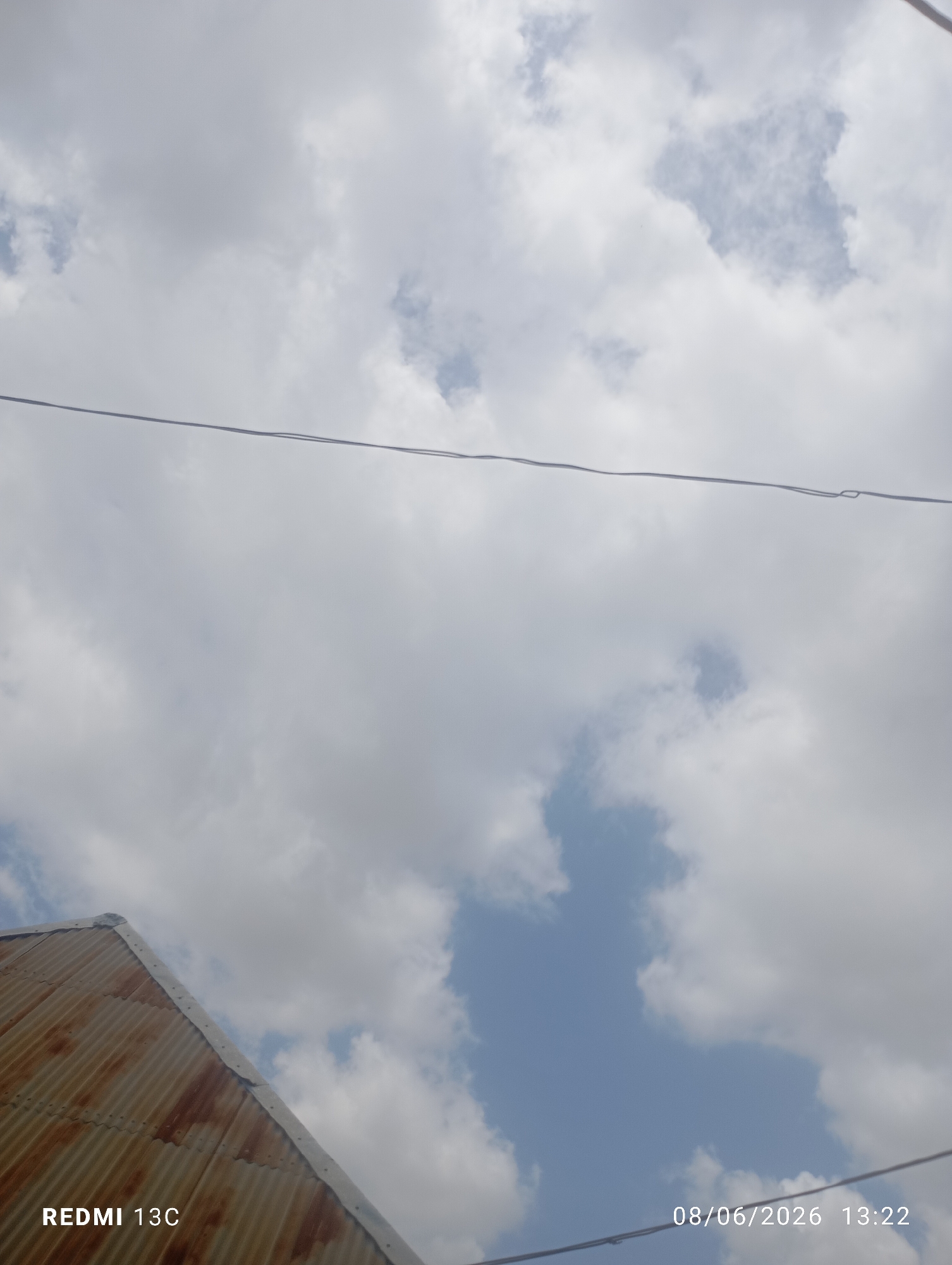 The image captures a cloudy sky with patches of blue visible. A portion of a rusty, corrugated metal roof is visible in the lower left corner, extending diagonally upwards. A thin wire runs horizontally across the middle of the frame. Text overlay in the lower left reads "REDMI 13C", and in the lower right reads "08/06/2026 13:22". The scene does not provide any identifiable landmarks or features to indicate a specific location within the city of Jos, Nigeria.