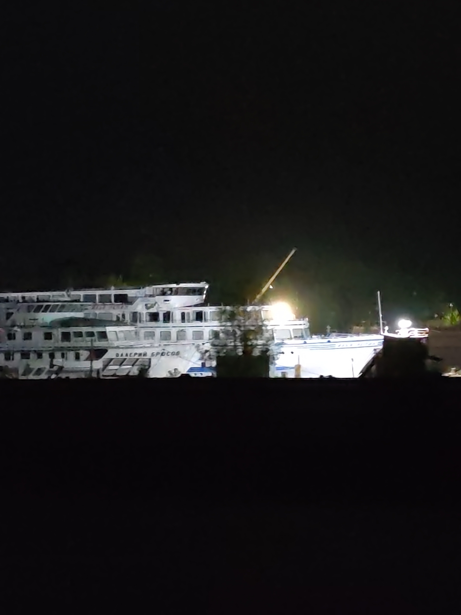 At night in Kimry, Russia, two docked vessels are observed. One is a three-tiered river cruise ship with the name "VALERIY BRUSOV" visible on its hull. A crane is positioned near the second vessel, a smaller, brightly lit boat. Both vessels are alongside a dark shoreline. The overall lighting is subdued, with the vessels illuminated against a dark background.