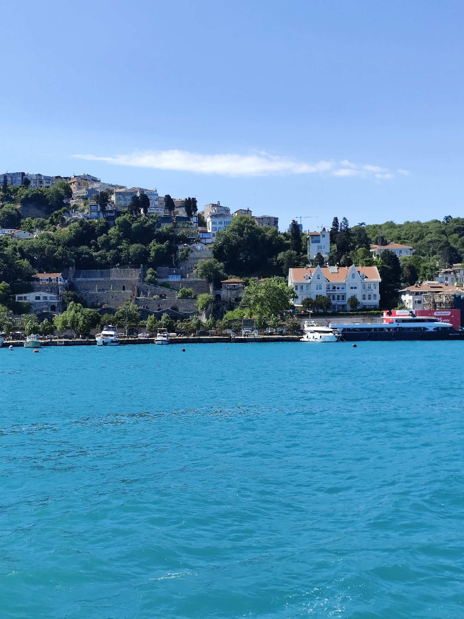 The image depicts a scenic view of the Bosphorus Strait in Beşiktaş, Turkey. The foreground is dominated by the turquoise water with gentle ripples. Along the shoreline, various structures are visible, including white and off-white buildings, with one prominent building featuring a red-tiled roof. A large yacht is docked near the shore. Behind the waterfront, a hillside rises steeply, covered with lush green trees interspersed with residential buildings, giving the landscape a layered appearance. The sky is blue with a few wispy clouds. A portion of Rumeli Hisarı is visible on the hillside.
