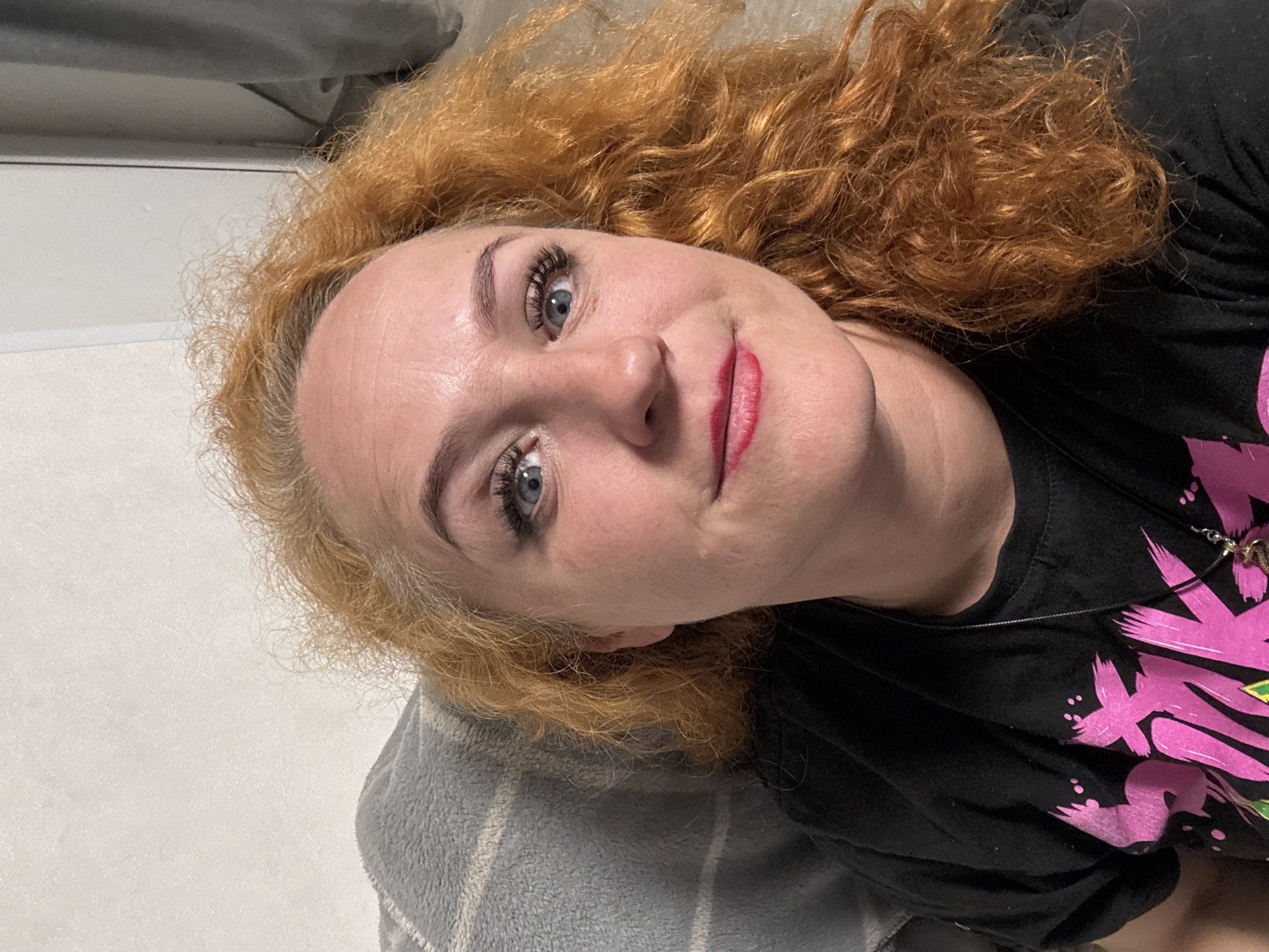 A woman with fair skin, red curly hair, and blue eyes is lying down. She's wearing a black t-shirt with a pink design. Her head is resting on a gray and white striped pillow. She's wearing makeup, including lipstick and mascara. The background is plain and out of focus. The image appears to have been taken indoors. There is no evidence that the image was taken in Chaikovsky, Russia.