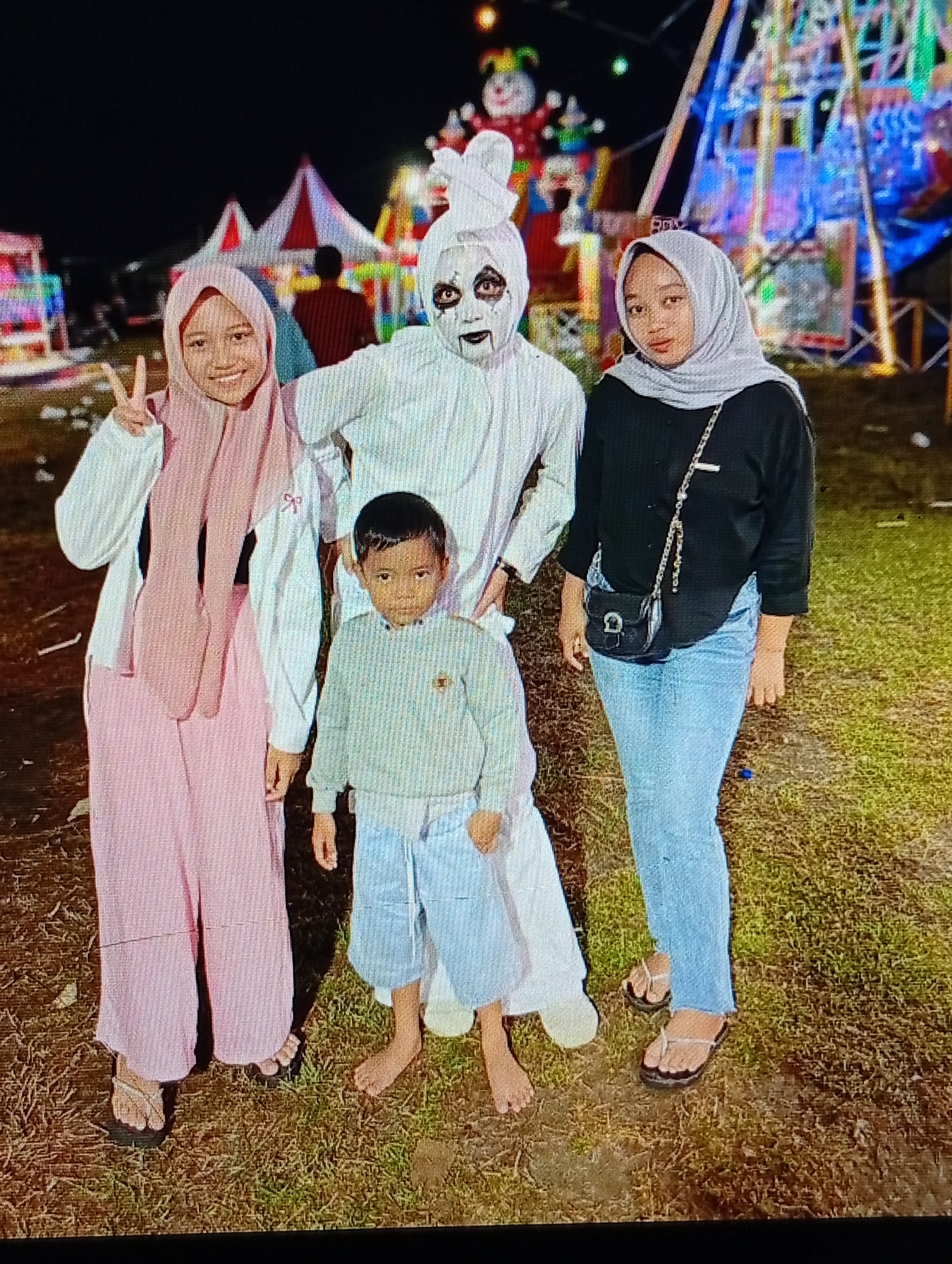 A nighttime photograph taken in Lhokseumawe, Indonesia, depicts four individuals posing at what appears to be a fairground or carnival. On the left, a young adult female wears a pink hijab, a white long-sleeved cardigan, light pink wide-leg pants, and dark flip-flops; she smiles and makes a peace sign with her right hand. In the center, an adult figure is dressed in a white, full-body "pocong" ghost costume, featuring white fabric wraps on the head, black makeup around the eyes, and dark lips; this individual stands with hands on hips. A young male child, estimated to be between 4-7 years old, stands slightly in front of the ghost figure, wearing a light blue long-sleeved top and light blue shorts, and is barefoot. On the right, a second young adult female wears a grey hijab, a black long-sleeved top, light blue jeans, and light-colored flip-flops, with a small black cross-body bag slung over her shoulder. All subjects face the camera directly.
The background, set against a dark night sky, features various brightly lit amusement attractions. To the far left, colorful illuminated stalls are visible. Two prominent red and white striped tents stand behind the subjects, with a blurred figure walking away in the distance behind them. Further to the right, a large, brightly lit amusement ride, possibly a Ferris wheel or similar structure, glows with blue and white lights. A large, illuminated clown-like character with a red nose and green hat is visible behind the ghost figure. The ground beneath the subjects is a mixture of dry, patchy grass and dirt, with some small debris scattered. Artificial lighting from the carnival rides illuminates the scene, casting soft shadows.
