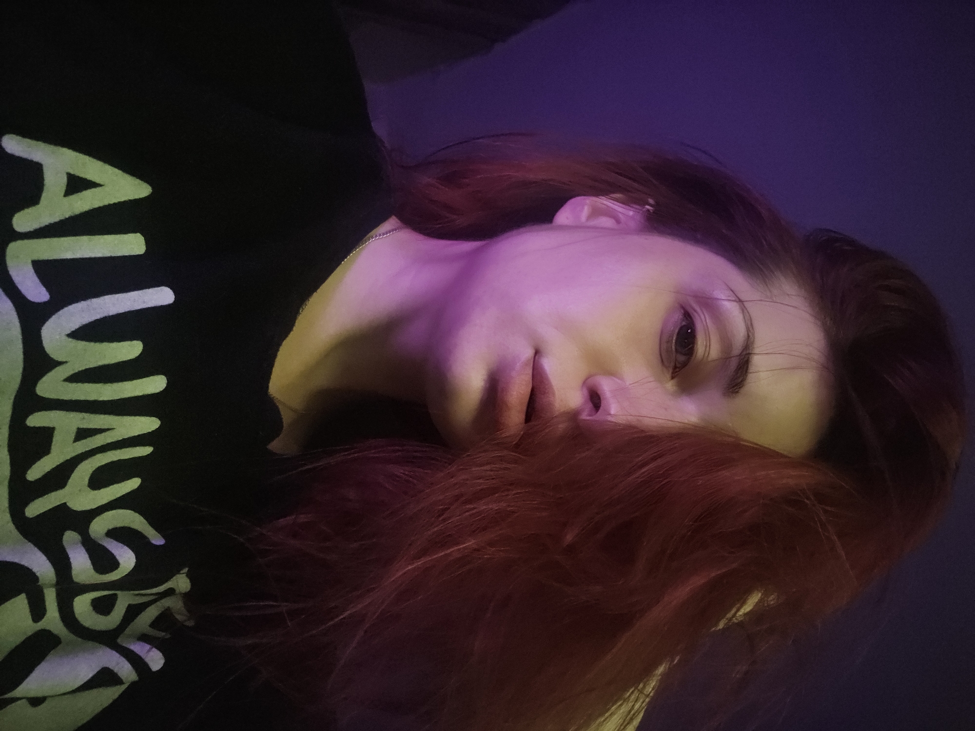 A close-up, overhead, indoor, selfie-style photograph shows a young adult, female, with fair skin, dark eyebrows, and bright red, somewhat messy, hair. She is wearing a black t-shirt with white text that reads "ALWAYS BE". The background is a gradient of dark purple to dark blue, suggesting an artificial light source. The subject's head is tilted, and her hair is falling across the right side of her face, partially obscuring her eye. Her lips are slightly parted, and she appears to be looking directly at the camera. The lighting casts a pinkish-purple hue on the left side of her face and neck. No specific location in Burgas, Bulgaria can be identified from the image.