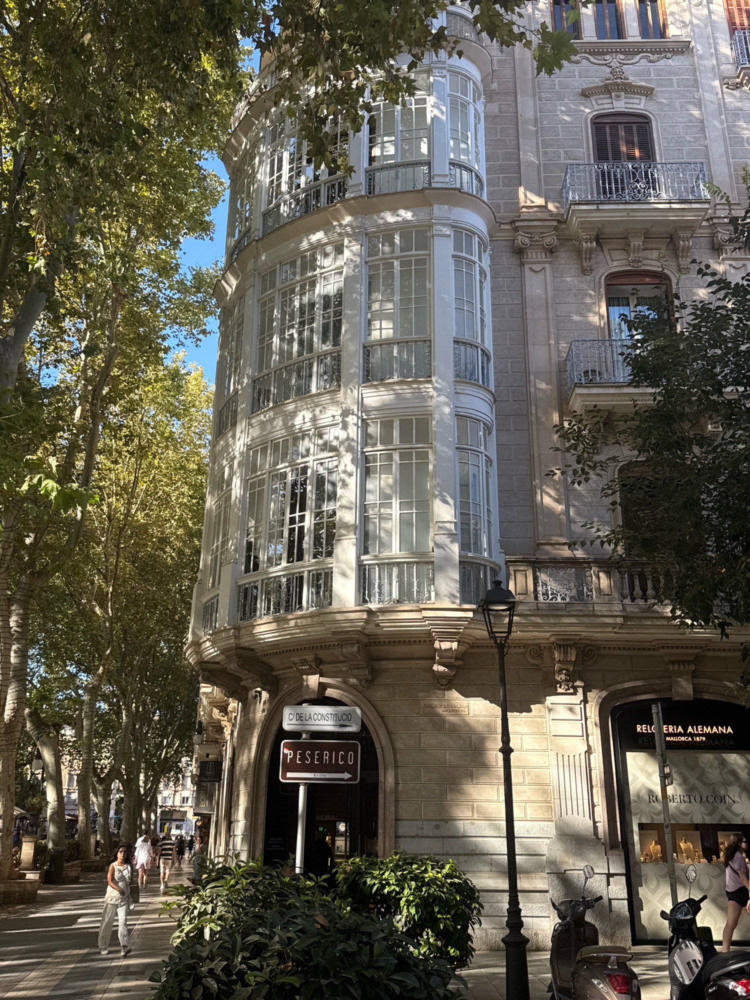 The image captures a sun-drenched street scene in Palma, Spain, featuring a grand architectural building with ornate balconies and bay windows on the corner of a street lined with mature trees. A street lamp stands prominently in the foreground. A sign above an arched entrance reads "C/ DE LA CONSTITUCION" and another below it points to "PESERICO". Further along the building, the signage for "RELICERIA ALEMANA MALLORCA 1879" and "ROBERTO COIN" is visible, indicating a jewelry store. Several people are walking along the sidewalk, and two scooters are parked near the building. The architecture suggests a historic European city, consistent with Palma de Mallorca.