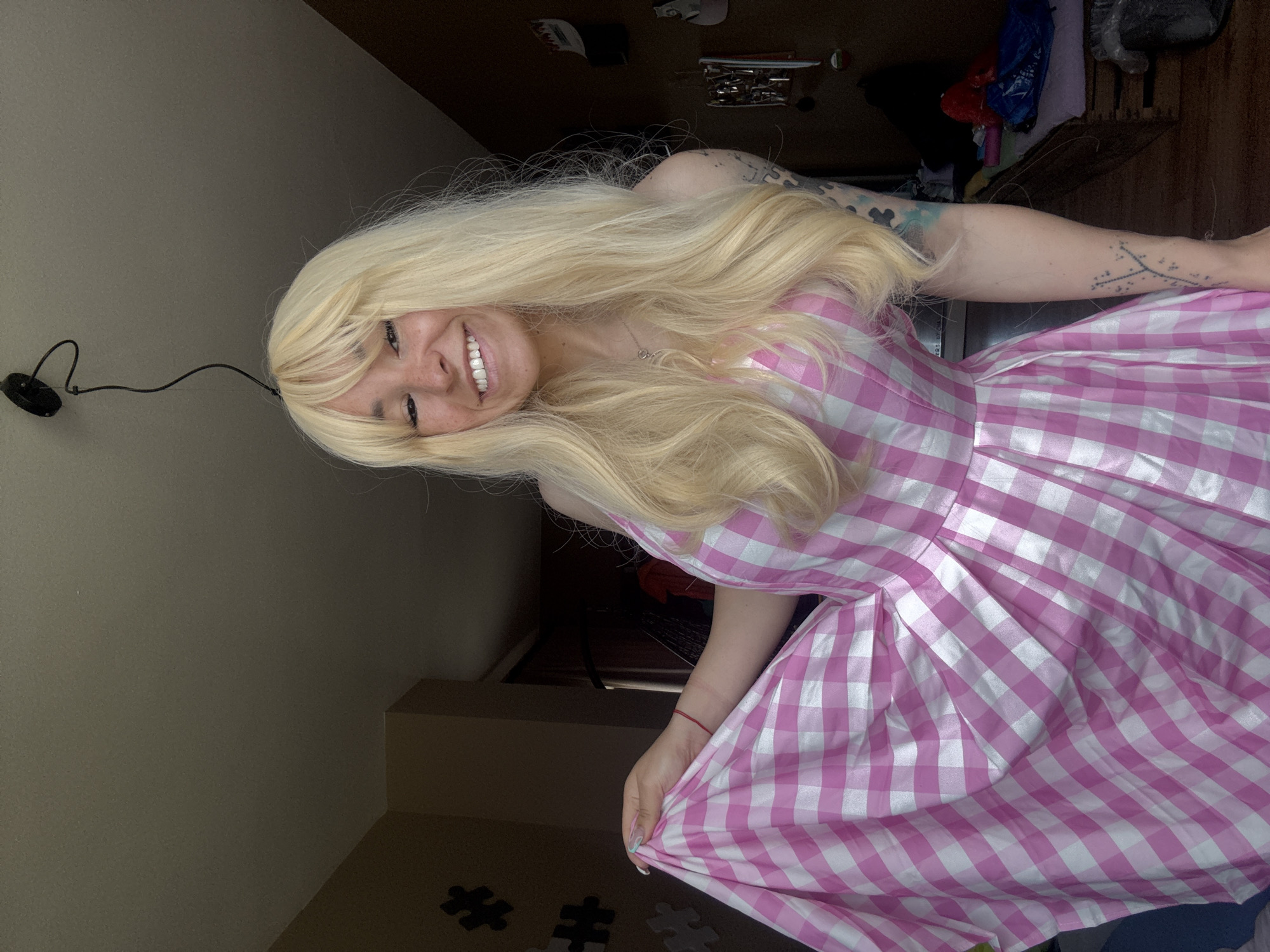 A young woman with blond hair and a pink and white checkered dress is smiling and looking at the camera. Her dress is full and gathered at the waist. She has tattoos on her left arm. She is indoors. The location is not discernible from the image.