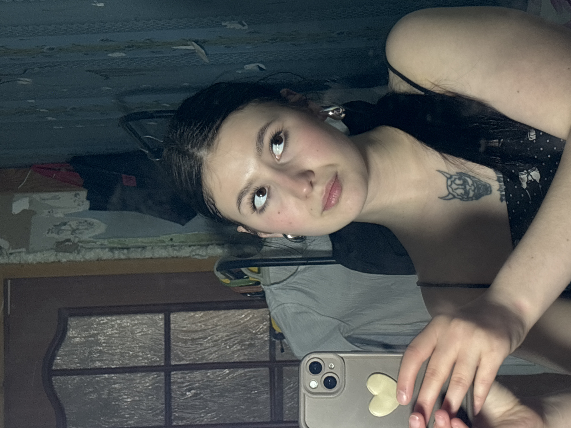 A young woman is lying down, taking a selfie in what appears to be a room with a dated interior. She has dark hair, fair skin, and is wearing a black top. A tattoo is visible on her chest. The background includes a patterned ceiling, and a reflective surface shows a windowed door and other room contents. The photograph does not contain any information about the location of Фоки, Russia.
