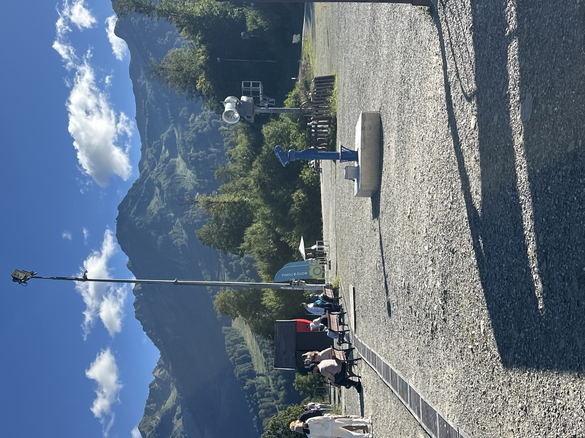 The image depicts a sunny day in Estosadok, Russia, with a mountainous backdrop and a paved area in the foreground. A tall metal pole with lights and a speaker system stands prominently, casting a long shadow across the gravelly ground. In the middle ground, several people are seated on benches, some interacting with each other, while others appear to be waiting. A small red building with a dark roof is visible behind the benches. A blue telescope is mounted on a concrete base in the foreground, pointing towards the mountains. A blue flag with text is planted to the right of the building. The environment is a mix of natural beauty with lush green mountains and man-made structures, suggesting a tourist or recreational area. The overall scene is peaceful and offers a view of the surrounding landscape.