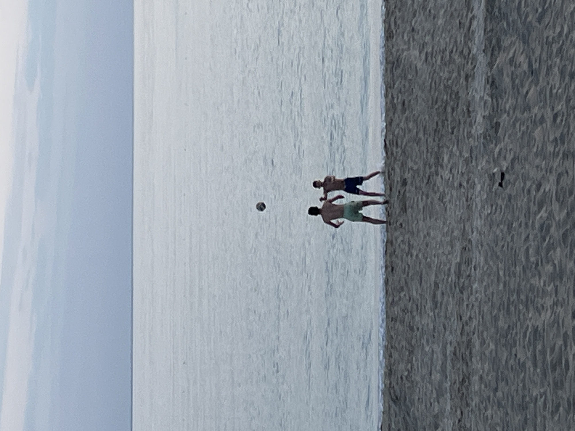 Two individuals stand on a beach playing with a ball. To the left is a body of water, and on the right, the beach continues. It's difficult to confirm the precise location in Alanya, Turkey without additional information.