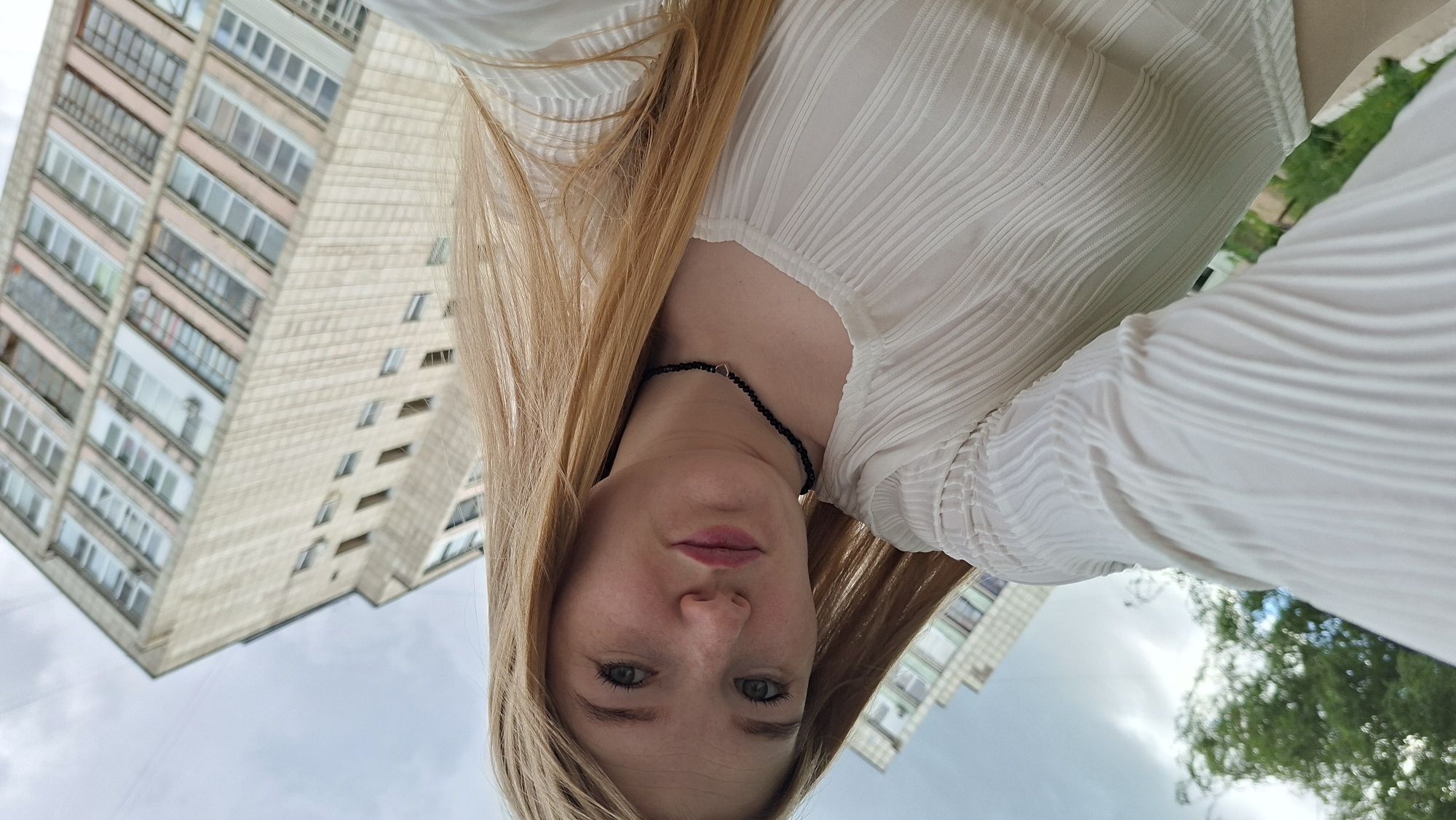 The image is a low-angle, wide shot of a young Caucasian woman with long blonde hair, wearing a white pleated top and a black beaded necklace. She is looking directly at the camera with her lips slightly parted. Behind her, a tall, beige apartment building with many windows is visible, angled upwards due to the camera's perspective. To the left, a blurry green tree and the sky with some clouds are present. The scene appears to be outdoors in an urban environment, likely Chaikovsky, Russia, given the architectural style of the building.