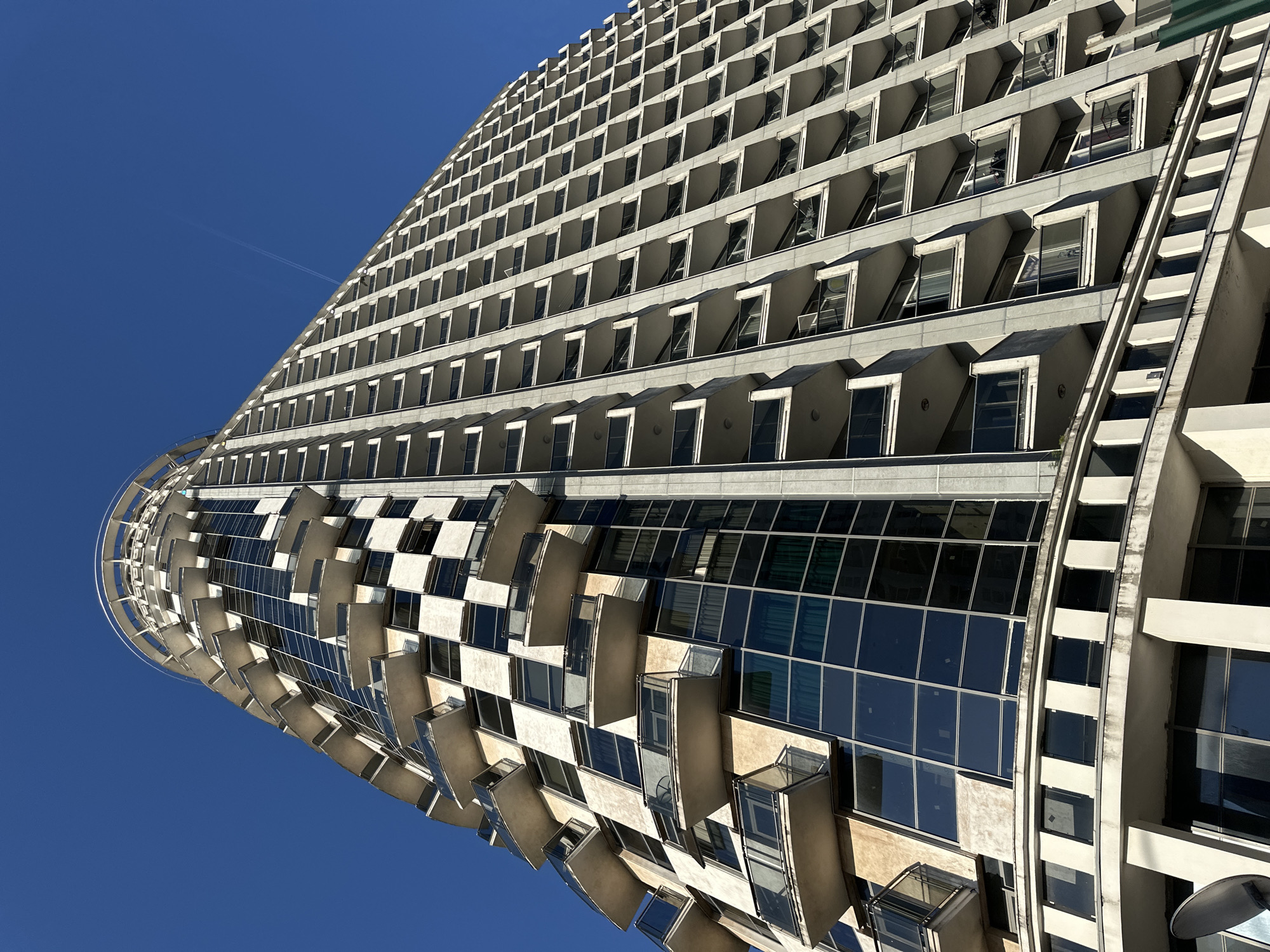 The image shows an upward-angled view of a tall, modern building against a clear blue sky. The building has a curved architectural design, featuring numerous rows of identical windows. The windows are recessed and have projecting elements above them, creating a layered effect. Some windows are open, revealing glimpses of the interior. The lower section of the building has a different design, with a checkerboard pattern of glazed and solid surfaces. The overall style suggests a modernist or brutalist architectural influence. The location is likely Batumi, Georgia.