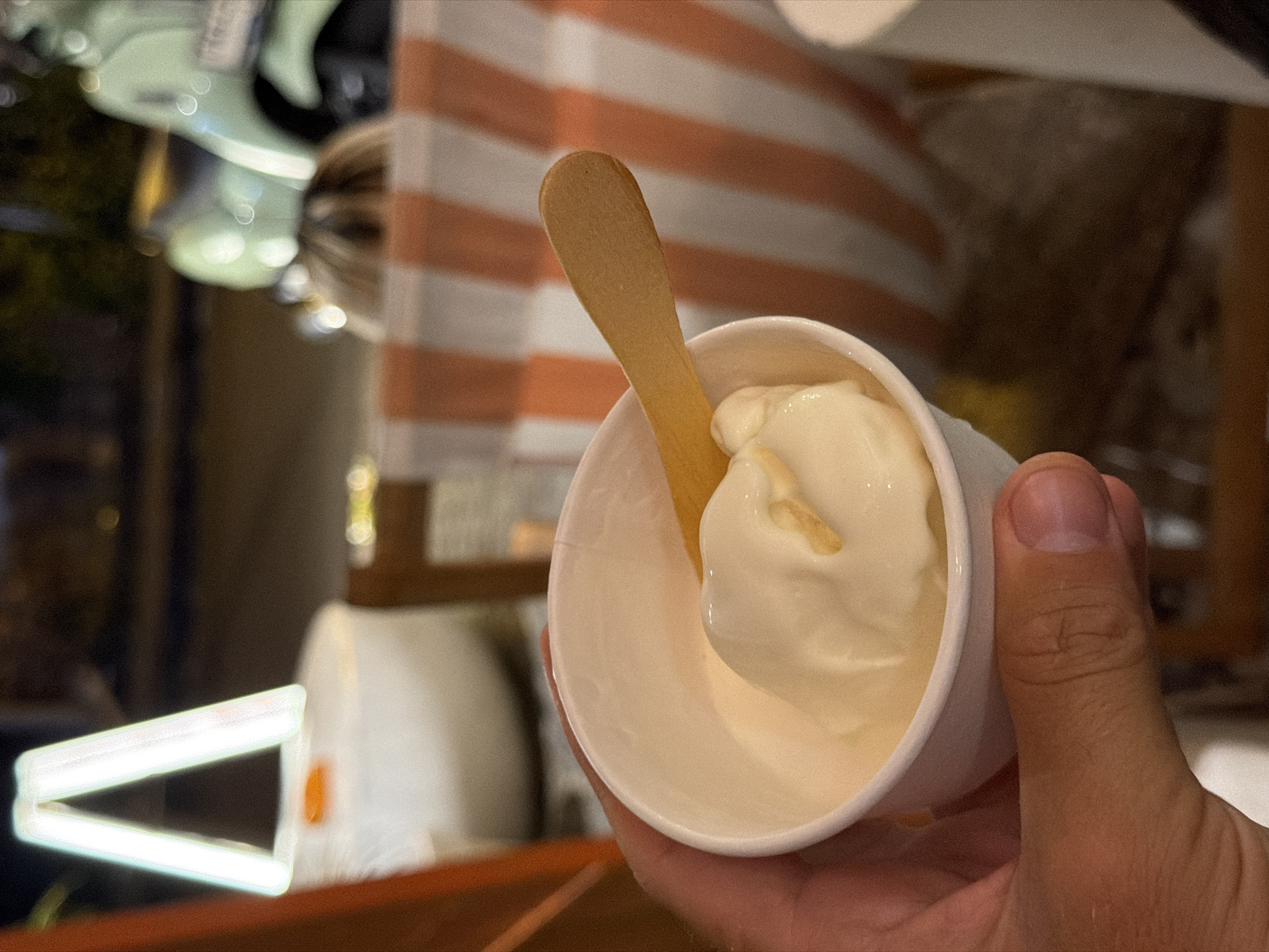 A hand holds a small white paper cup filled with white ice cream and a wooden spoon. Behind the cup, there is a background with vertical orange and white stripes. To the left, an illuminated white "V" shape is visible in a darker, blurred background, suggesting a building or structure. The photo was taken in the city of North Kuta, Indonesia.