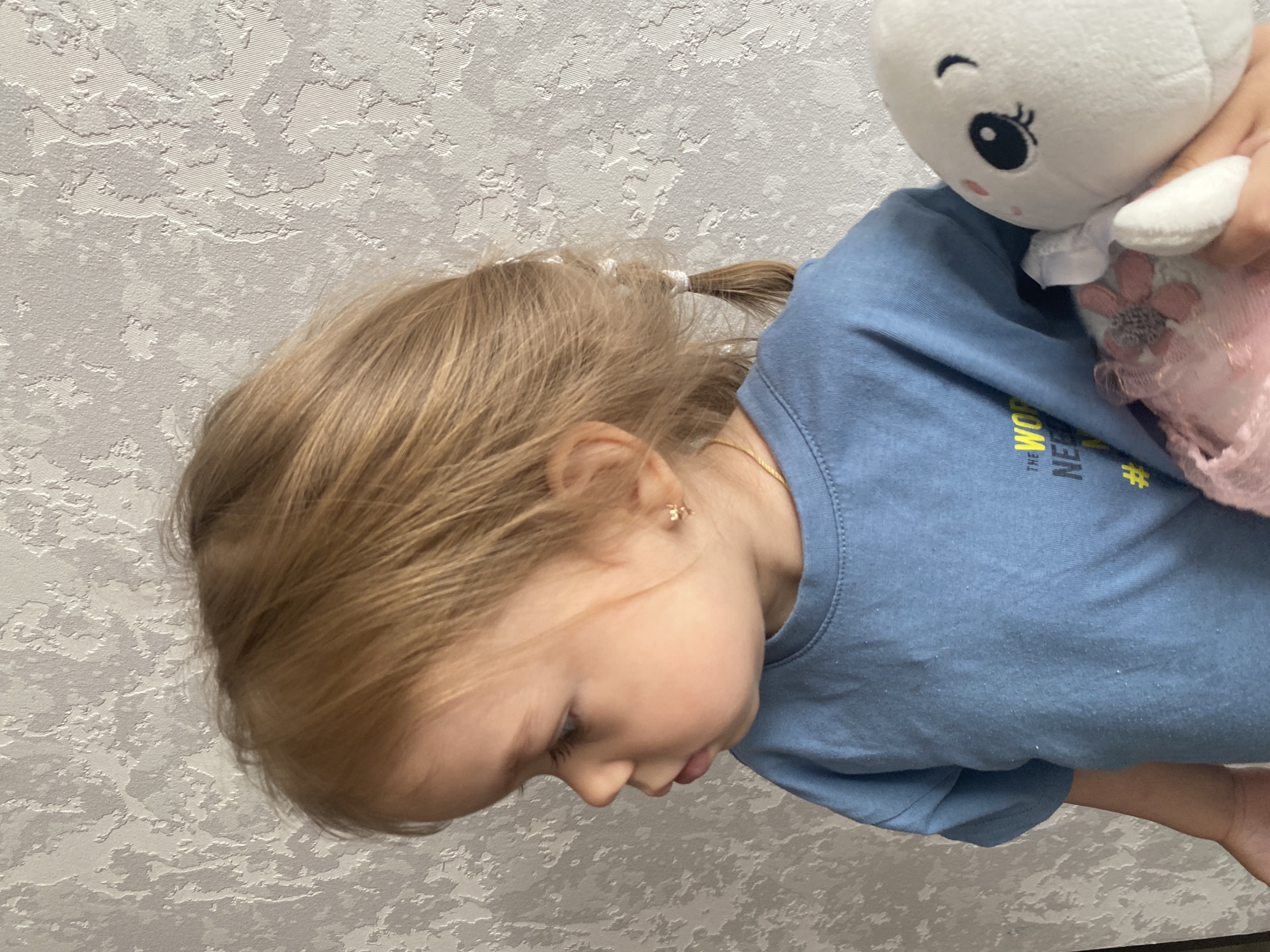 A blonde-haired child, wearing a blue t-shirt, is holding a stuffed animal against a textured, light-colored wall. The child's hair is pulled back into two pigtails, and they are wearing a small gold earring shaped like a star. The stuffed animal is white with black eyes and a pink blush on its cheeks. The background wall has a subtle patterned texture. The image appears to be taken indoors, possibly in a home. There is no discernible information that places this image specifically in Chaikovsky, Russia.