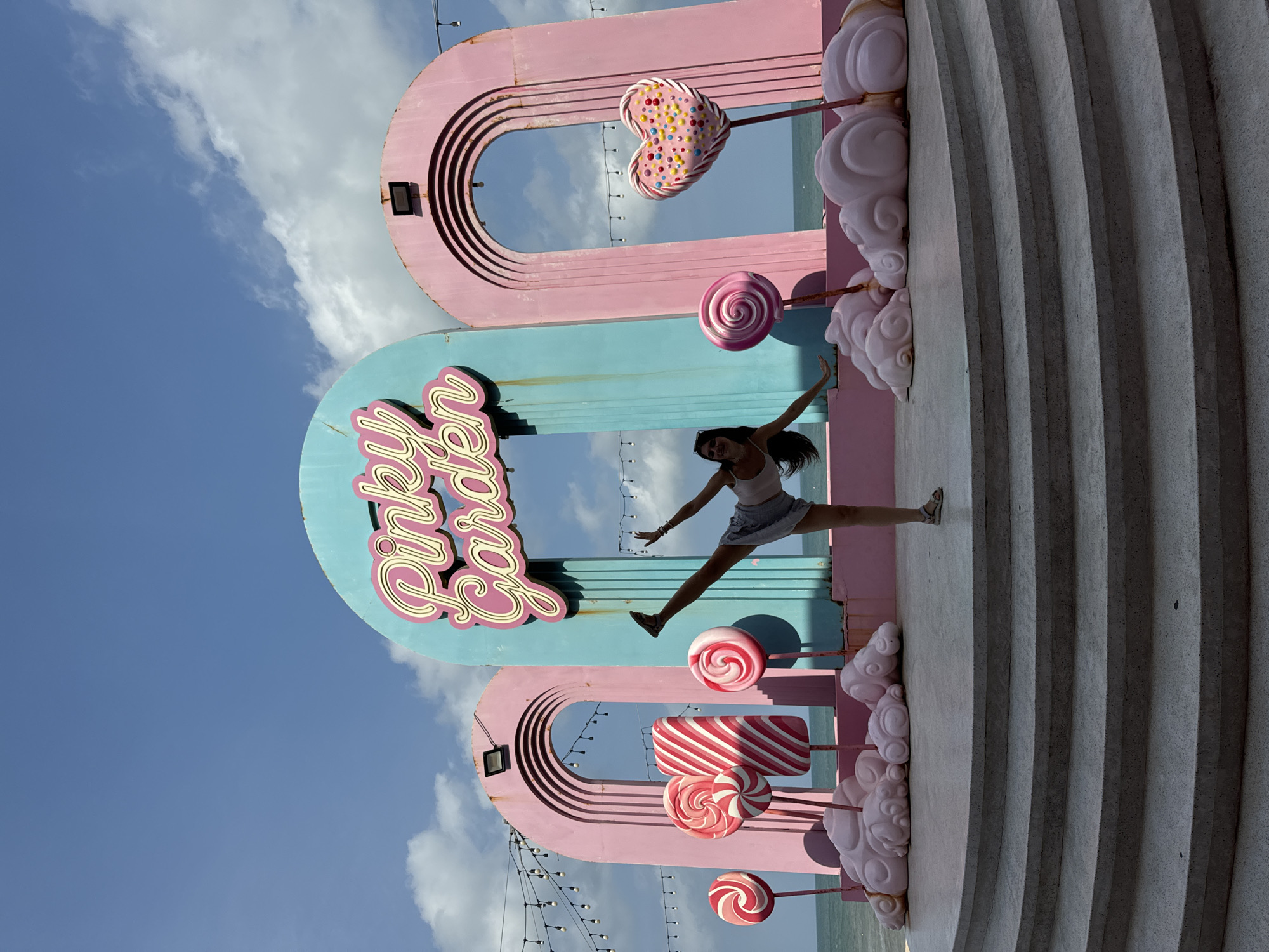 A young woman performs a high kick in front of a whimsical, candy-themed installation located at Pinky Garden in Tien Thanh Ward, Vietnam. The installation features large, pastel-colored archways, oversized lollipops, and cloud-like structures. The woman, dressed in a white tank top and grey shorts, is positioned centrally, her right leg extended upwards and her arms outstretched. Behind her, the turquoise and pink archways frame the blue sky and the ocean. String lights are visible along the top of the structure. The ground is a tiered, circular concrete platform.