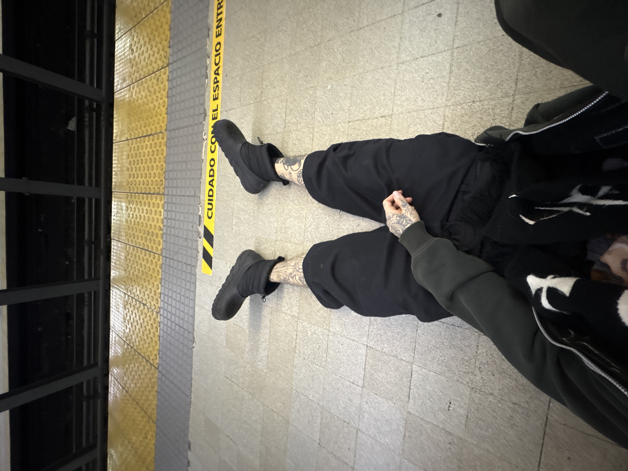 A person is seated on the floor of a subway platform, with their legs extended towards a safety strip. The strip reads "CUIDADO CON EL ESPACIO ENTRE". To the left of the safety strip is a yellow tactile paving surface, followed by the black frame of a subway car. The person is wearing black pants and shoes, with visible tattoos on their ankles and hands. They are also wearing a gray-green sweatshirt and a black and white jacket. Another person can be seen partially in the upper right corner, wearing black. The platform floor consists of light gray square tiles. The location is a subway station in Buenos Aires, Argentina, indicated by the Spanish language on the safety strip.