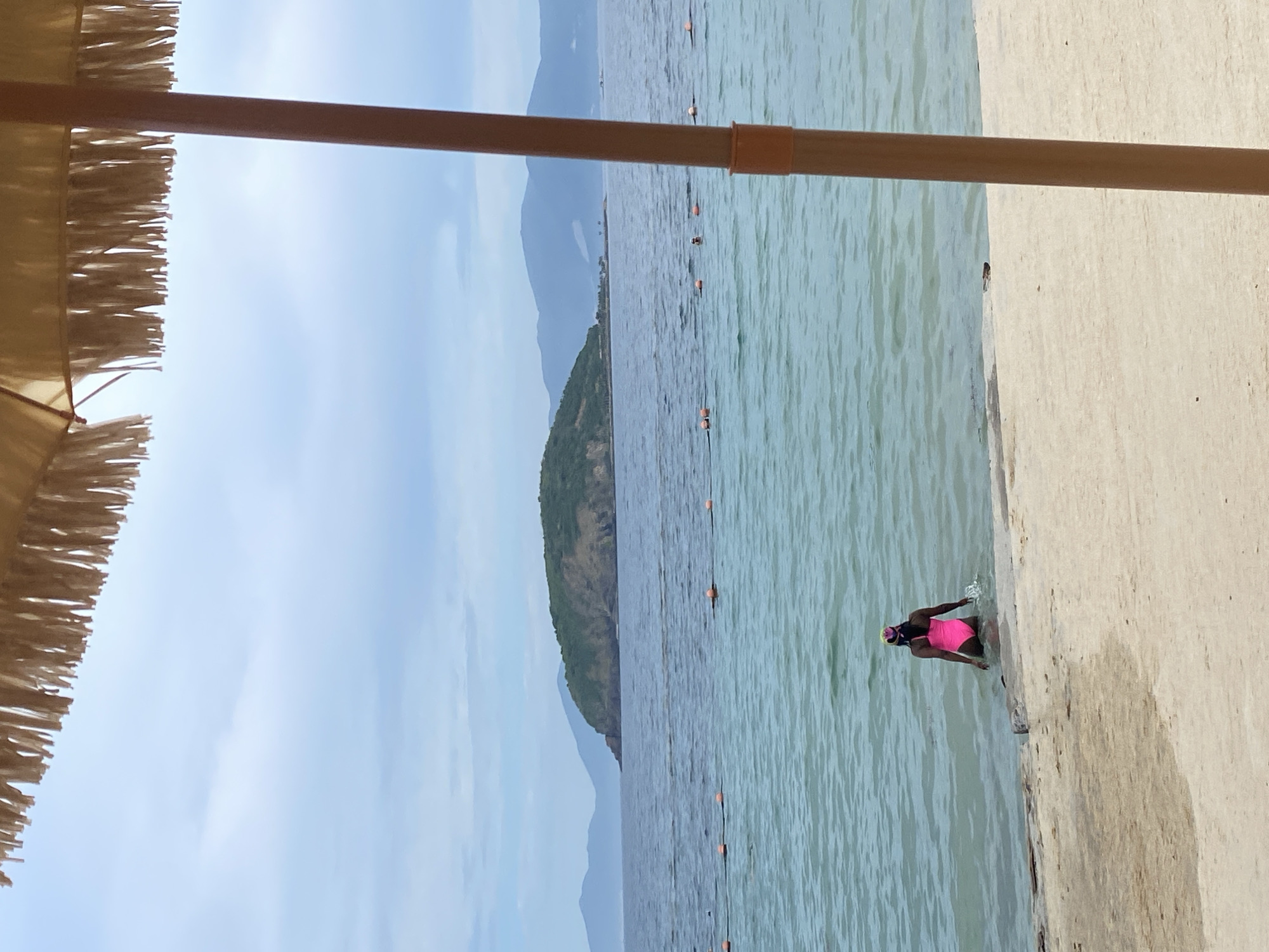 A person wearing a bright pink swimsuit and a colorful swim cap is standing at the edge of the water, facing away from the camera, on a sandy beach. The person is partially submerged in the turquoise water, with their legs and lower torso in the sea. The beach transitions to calm water, and in the distance, a lush green island is visible, surrounded by mountains under a cloudy sky. Orange buoys are spread across the water, indicating a designated swimming area. A portion of a large straw umbrella is in the foreground, partially obscuring the view of the beach and the sky. The scene appears to be a tranquil day at a coastal location, possibly in Nha Trang, Vietnam, known for its beautiful beaches and islands.