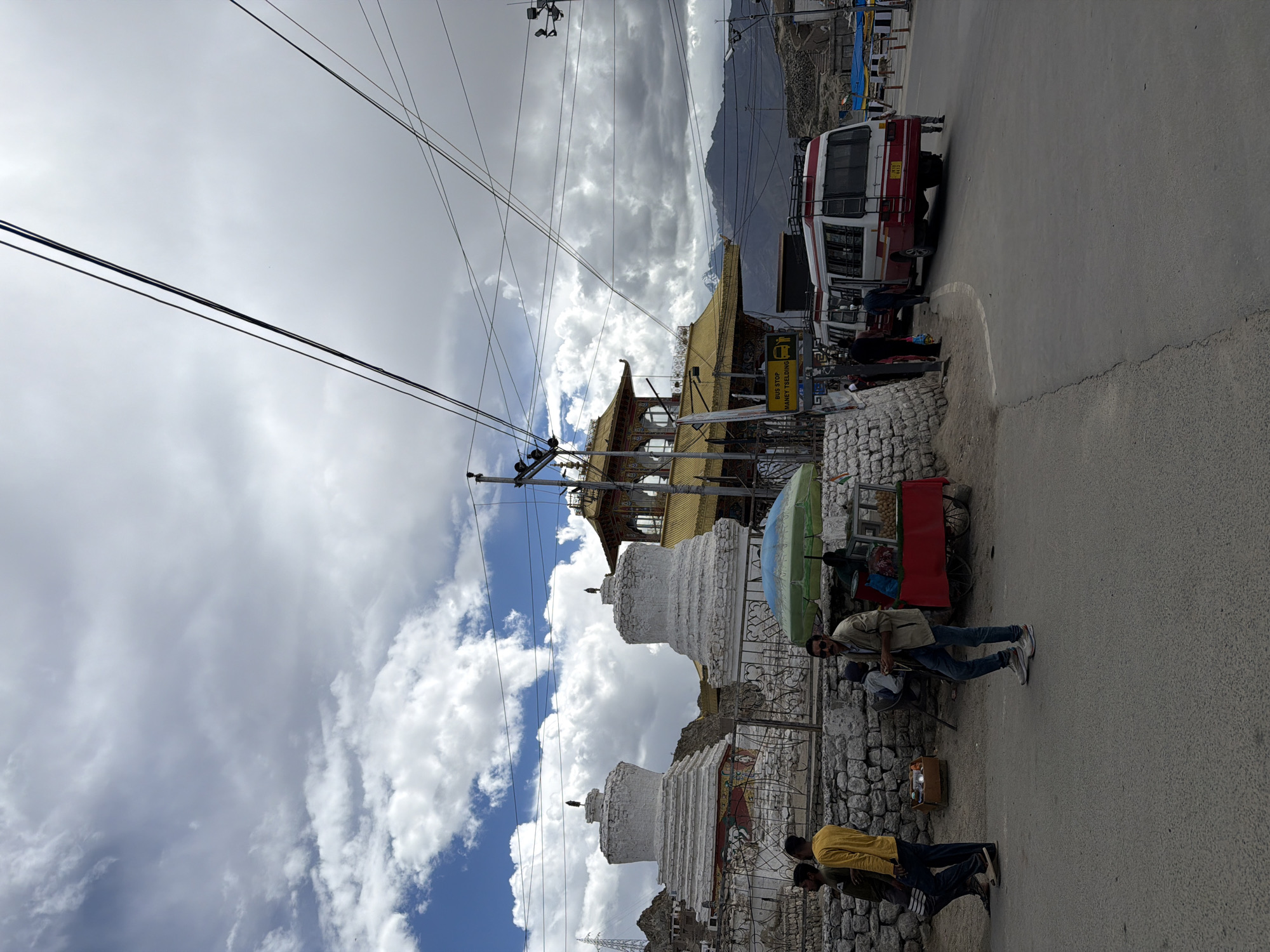 The image captures a street scene in Leh, India, near the Shanti Stupa or a similar Buddhist monument, as suggested by the prominent white stupas in the foreground and the temple-like building with a golden roof in the background. Several people are present: three men are walking on the left side of the street, one wearing a yellow shirt, another in a green shirt, and a third in dark clothing. A man in a light brown jacket and jeans stands in the middle of the road, looking towards the right. A food stall with a red base and a green umbrella is set up on the side of the street, with at least one vendor visible behind it. Two white and red buses are parked on the right side of the road. The sky is filled with dramatic clouds, and mountains are visible in the distance. The environment appears to be an urban or semi-urban area with paved roads, a stone wall, and overhead electrical wires.