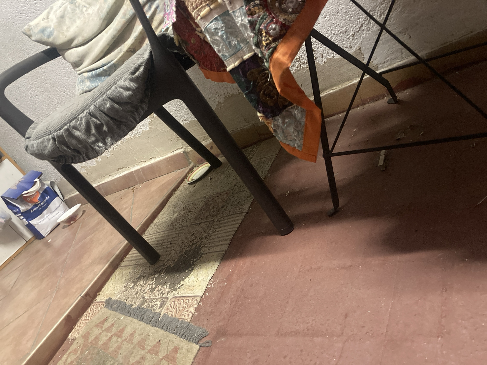 The image depicts an interior scene, likely a room with tiled flooring. A dark metal chair is positioned centrally, draped with various textiles. One textile is a gray corduroy seat cover, and another is a colorful, patchwork-style throw or blanket with orange trim. Behind the chair, a bag labeled "Arena Absorbente" is visible, suggesting it contains absorbent material, possibly for pet litter or spills. The floor appears to be a combination of reddish-brown tiles and a patterned rug that shows signs of wear or staining in one section. The wall behind the chair is textured and light-colored. The lighting in the scene is dim. The scene does not provide any specific indicators of its location in Sabadell, Spain, nor does it show any individuals or specific activities.